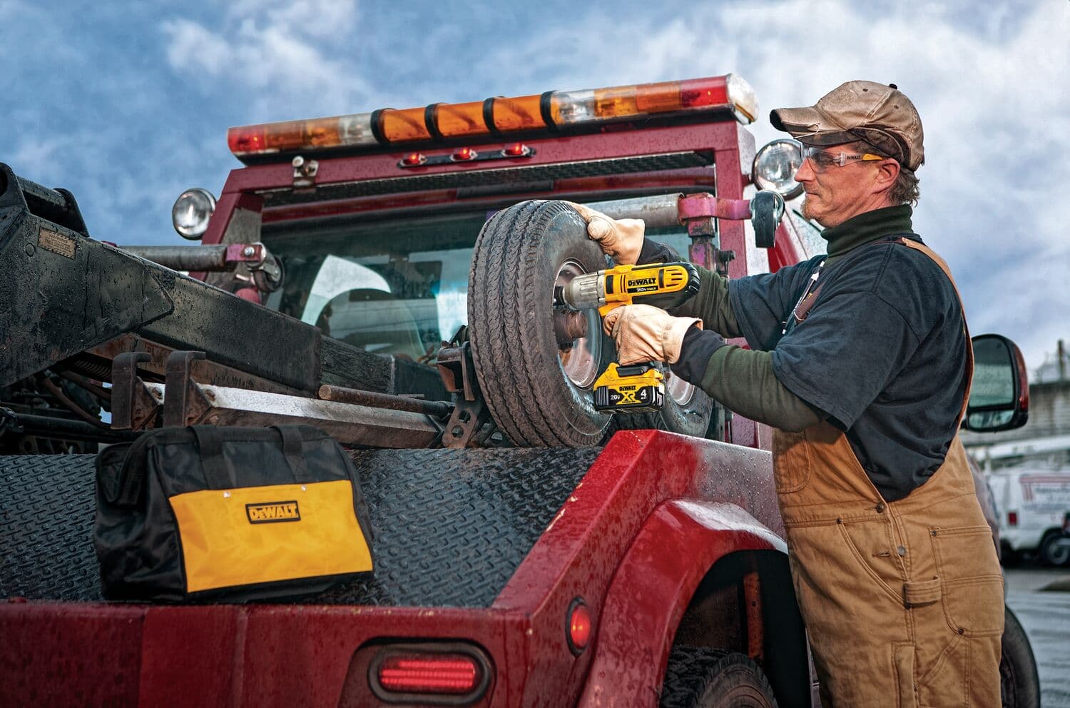 Una persona con un mono de trabajo está usando una llave de impacto inalámbrica amarilla DEWALT para quitar o apretar los pernos de una rueda de repuesto montada en la parte trasera de un camión utilitario rojo. Una bolsa de herramientas de la marca DEWALT está colocada en la cama del camión y el rostro de la persona está difuminado por privacidad.