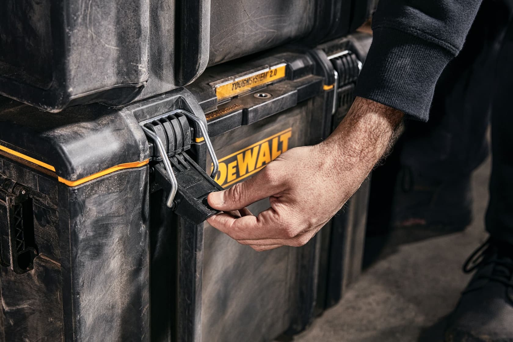 Primer plano de una persona preparándose para abrir el pestillo de una caja de almacenamiento DEWALT ToughSystem 2.0. La caja es negra con detalles amarillos y muestra signos de uso.