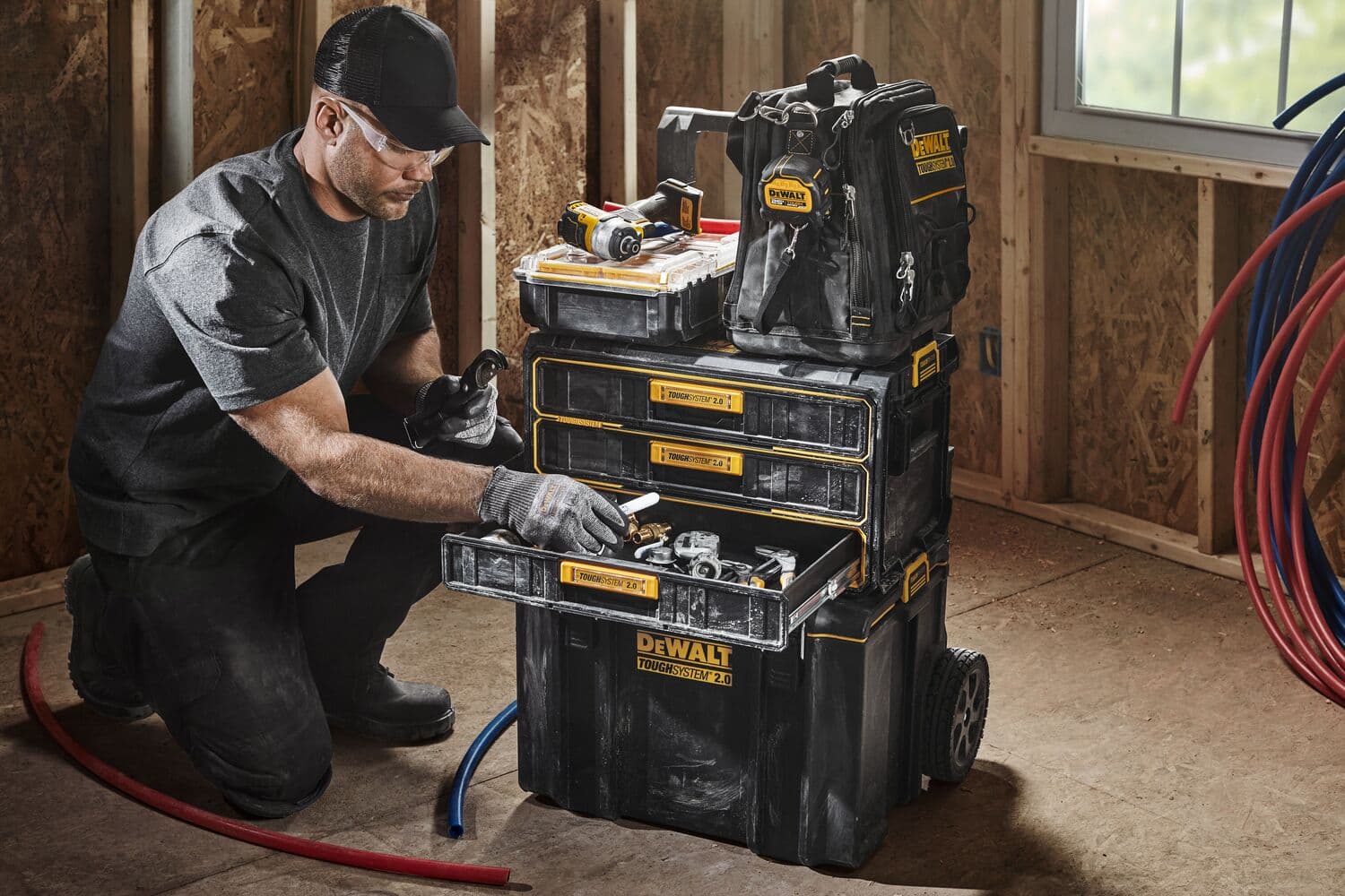 Un trabajador con guantes y gorra está arrodillado junto a un sistema de almacenamiento de herramientas apilable DEWALT ToughSystem 2.0. El sistema incluye varias cajas de herramientas negras y amarillas con cajones y compartimentos, llenas de diversas herramientas y accesorios. También hay una mochila DEWALT y herramientas eléctricas encima. La escena se desarrolla en una habitación sin terminar con paredes de madera y cables visibles.