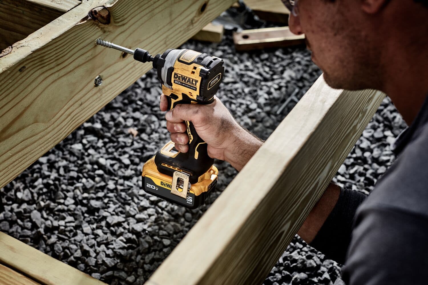 Una persona utiliza un atornillador de impacto inalámbrico DEWALT DCF850B para atornillar en una estructura de madera, con grava debajo.