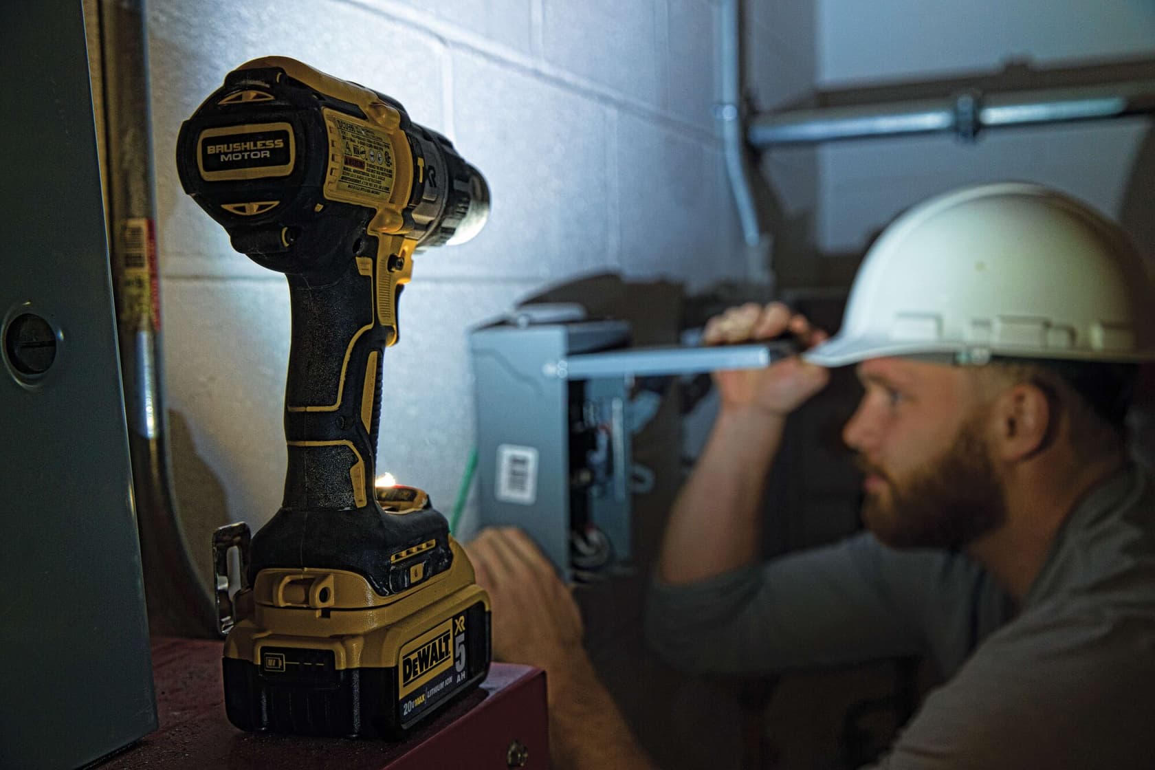 Un taladro inalámbrico DEWALT con motor sin escobillas está sobre una superficie en primer plano, mientras una persona con casco de seguridad trabaja en una caja eléctrica en el fondo. El rostro de la persona está difuminado para preservar la privacidad.