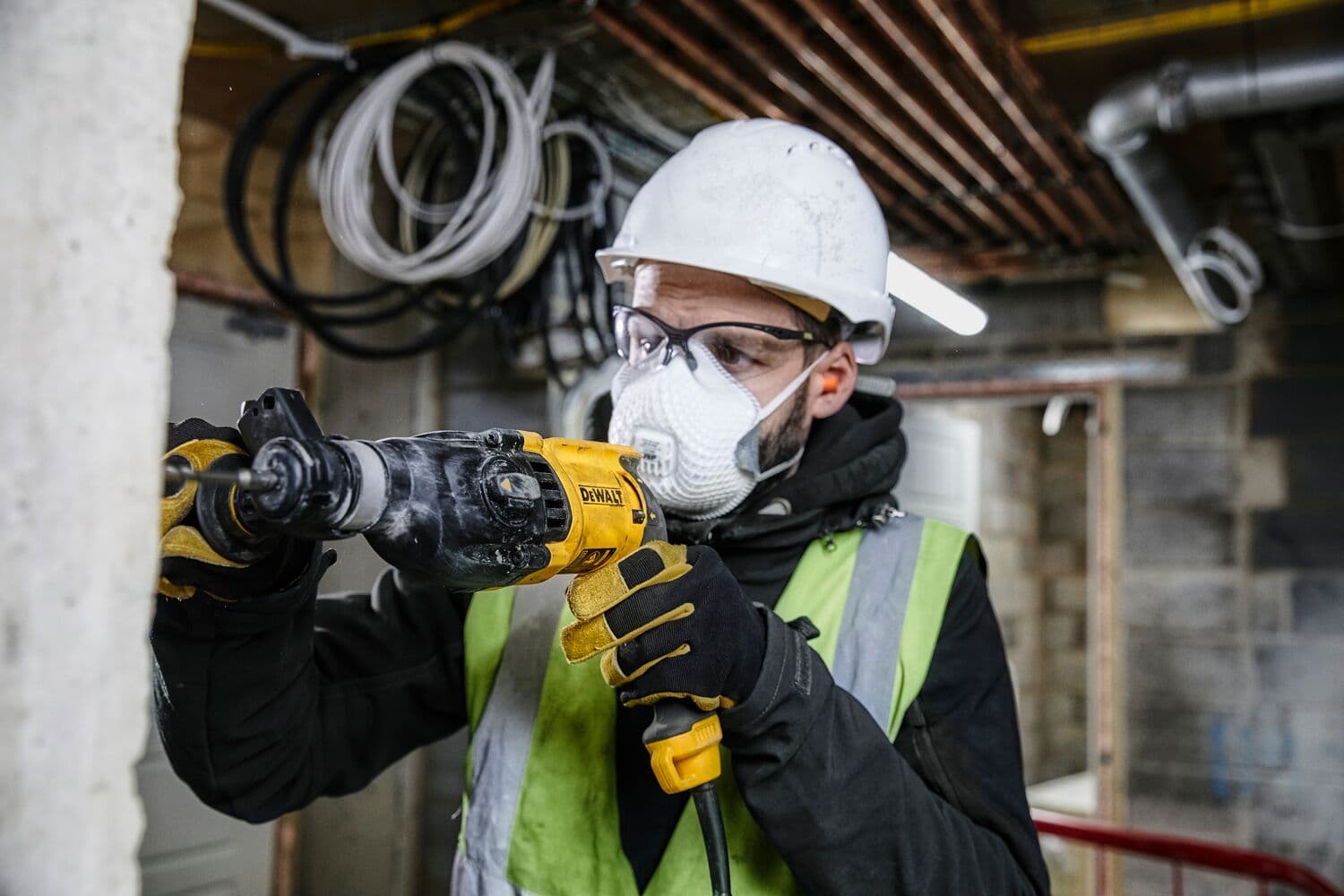 Una persona con casco de seguridad, guantes y chaleco reflectante está usando un taladro DEWALT para trabajar en una pared de hormigón en un entorno de construcción. El rostro está difuminado por privacidad.