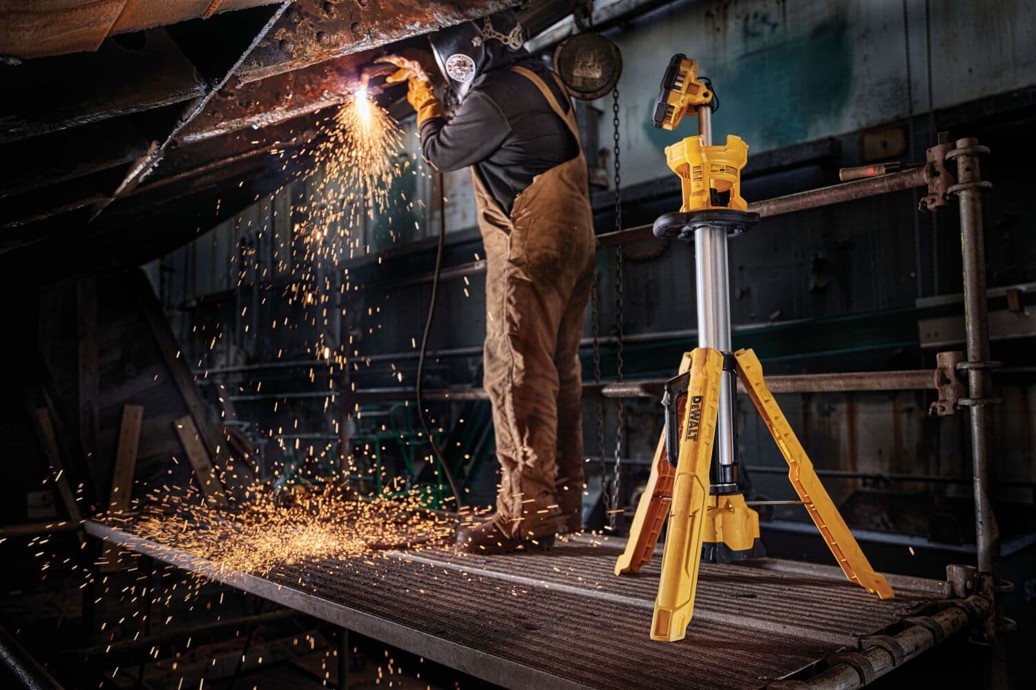 Una luz de trabajo con trípode DEWALT DCL079R1 está situada en una plataforma metálica cerca de una persona realizando soldadura, con chispas volando en un entorno industrial.