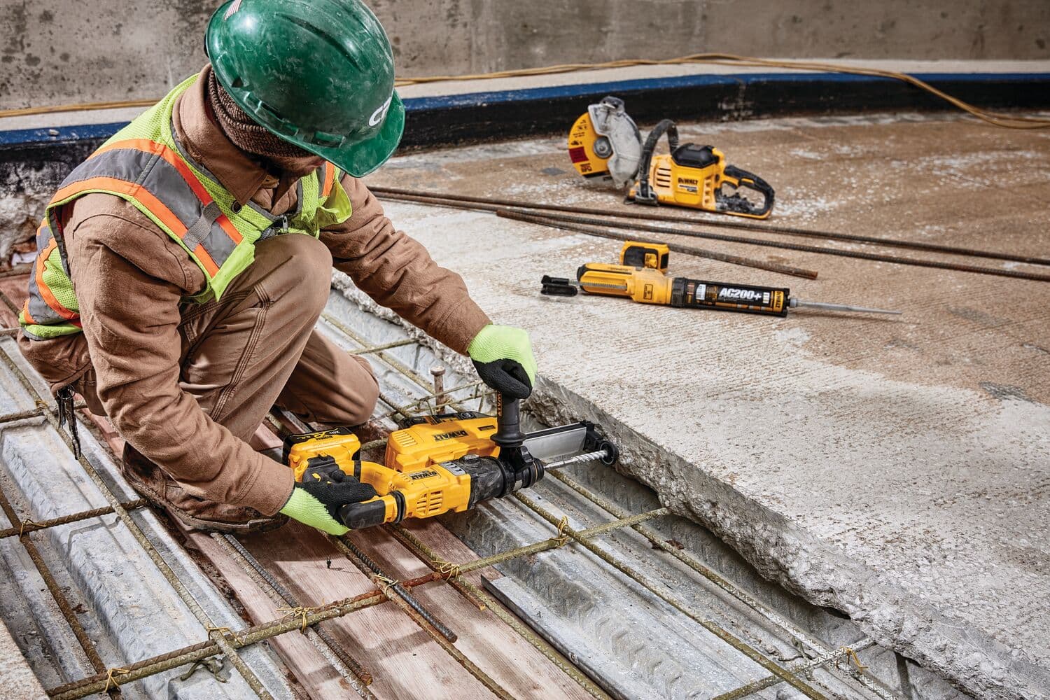 Un trabajador de construcción con equipo de seguridad y un casco verde utiliza una herramienta eléctrica amarilla DEWALT para perforar una losa de concreto. Más herramientas DEWALT, incluyendo una sierra y un martillo rotatorio, se ven en el suelo, todas destinadas a trabajos pesados de construcción.