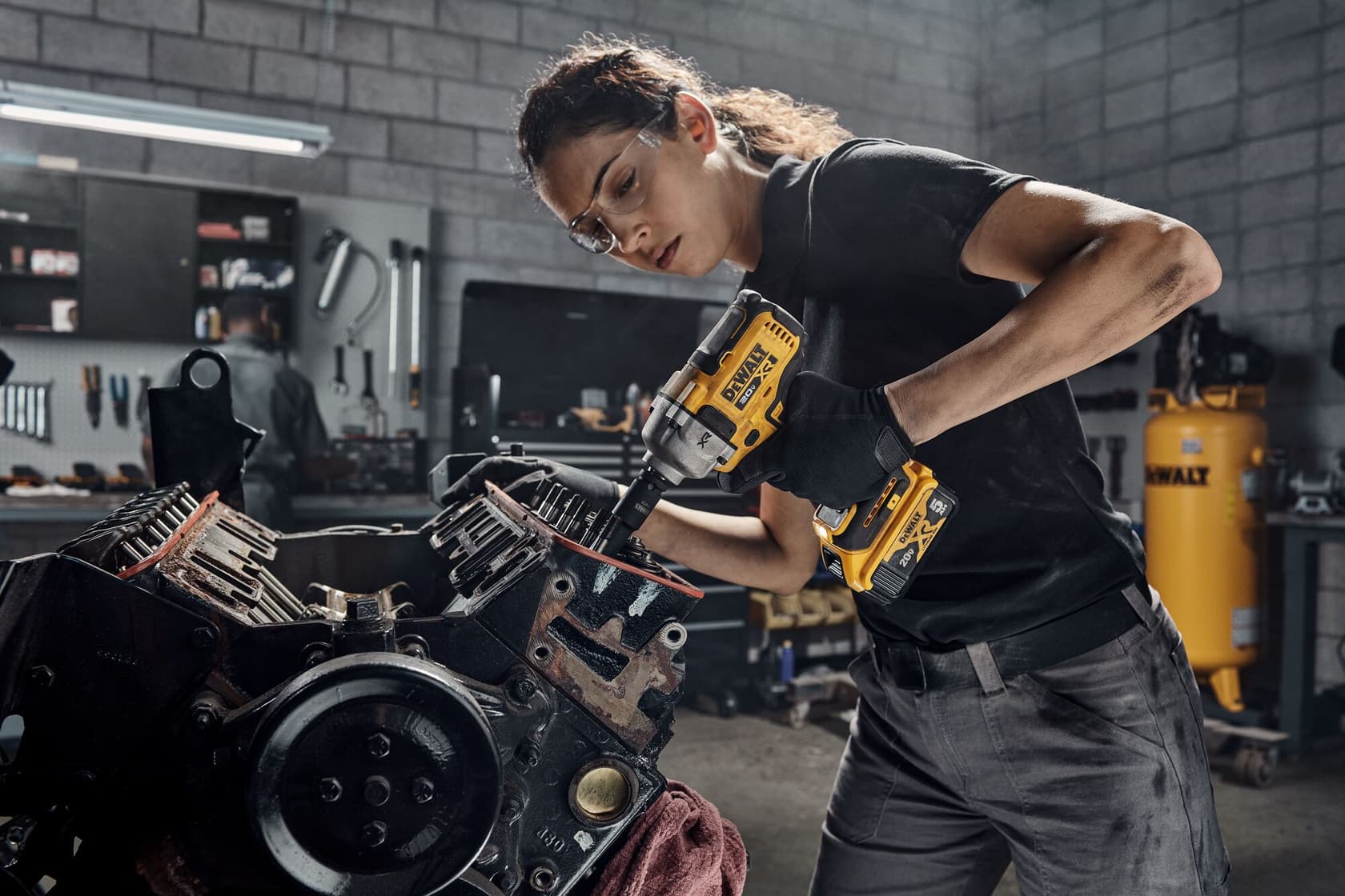 Una persona utiliza una llave de impacto inalámbrica DEWALT (modelo DCF892B) para trabajar en un motor en un taller. La cara de la persona está desenfocada por privacidad, y el fondo del taller incluye herramientas y equipos.