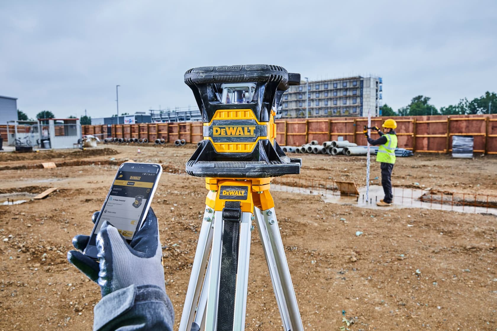 Nivel láser DEWALT sobre un trípode en un sitio de construcción, una mano enguantada sostiene un teléfono inteligente y un trabajador con chaleco de seguridad y casco mide en el fondo.