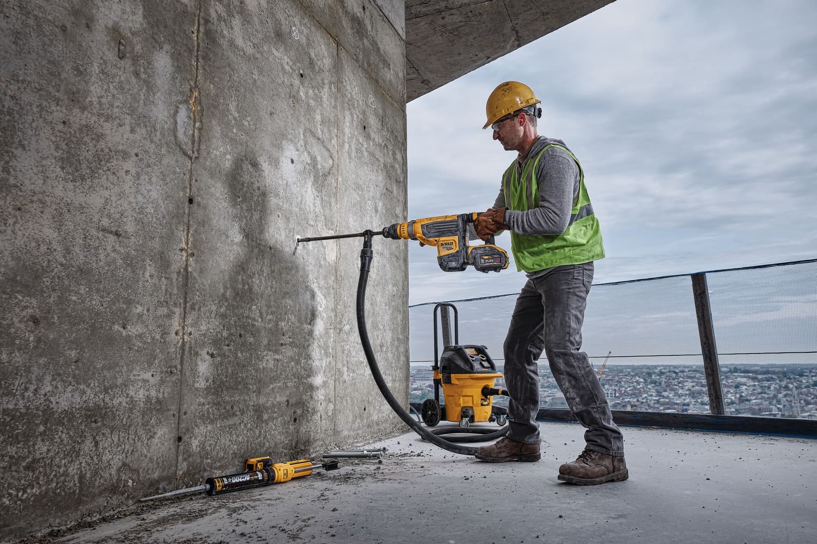 Un trabajador de construcción con casco amarillo y chaleco de seguridad verde utiliza un taladro rotativo DEWALT para perforar una pared de concreto. El taladro está conectado a un sistema de aspiración para reducir el polvo, y hay herramientas DEWALT adicionales sobre el suelo. La escena está al aire libre, en lo alto de una ciudad.
