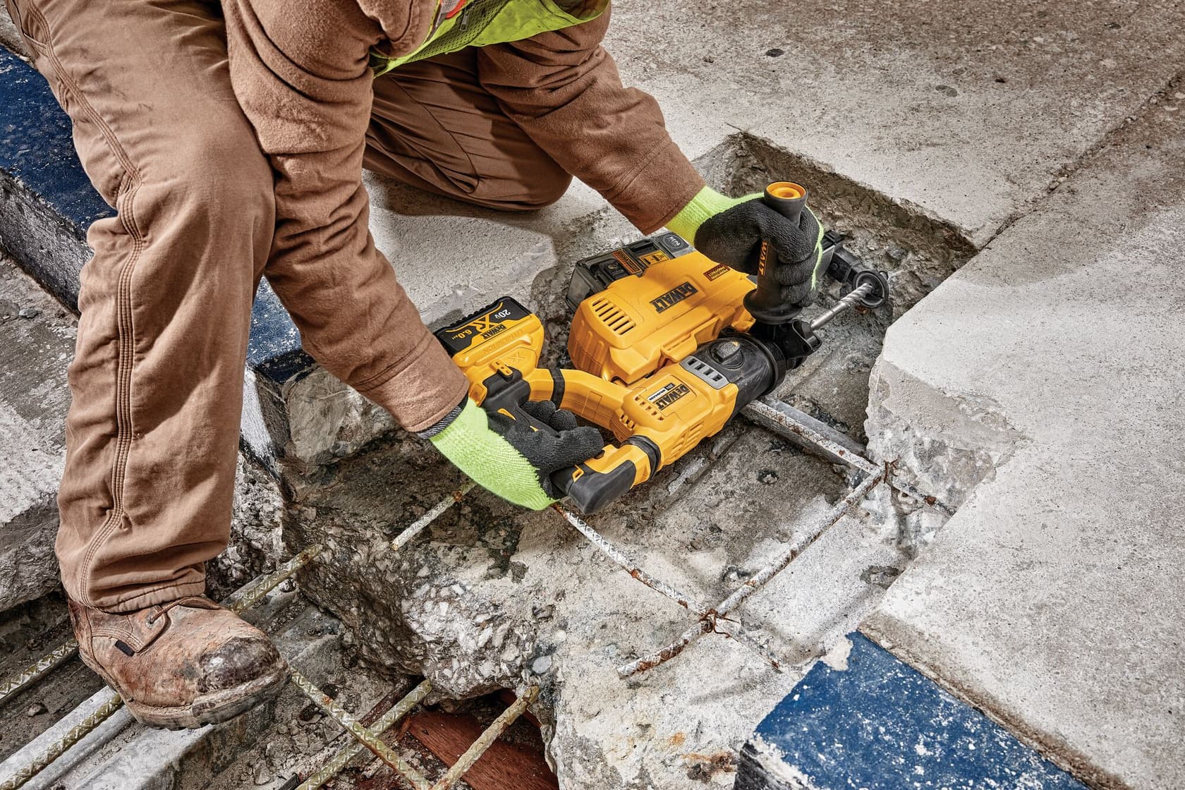 Un trabajador usa un martillo rotativo inalámbrico DEWALT con accesorio de extracción de polvo para perforar concreto, llevando guantes, botas de seguridad y pantalones de trabajo.