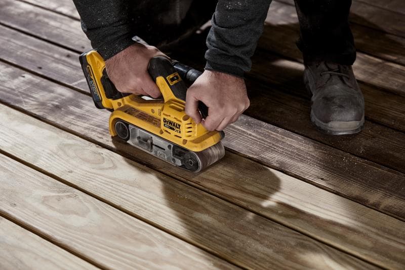 Una persona usa una lijadora de banda inalámbrica amarilla DEWALT para lijar tablas de madera en el suelo.