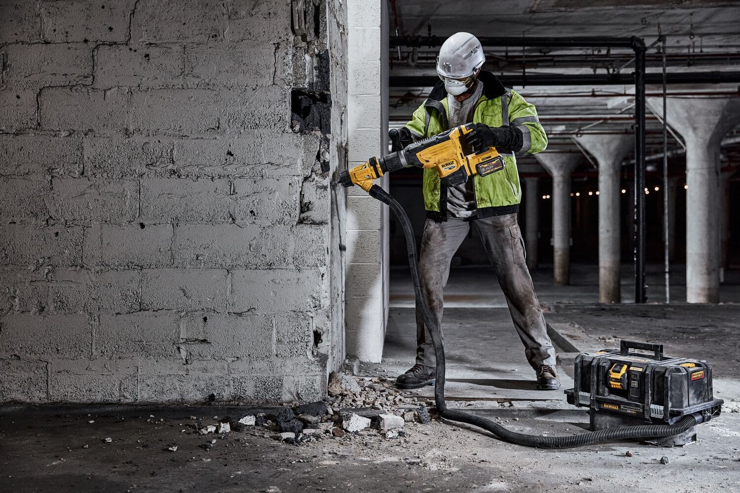 Un trabajador de la construcción con equipo de seguridad utiliza un martillo rotativo inalámbrico amarillo DEWALT en una pared de concreto. Hay escombros en el suelo y una caja de herramientas DEWALT con una manguera unida está cerca.