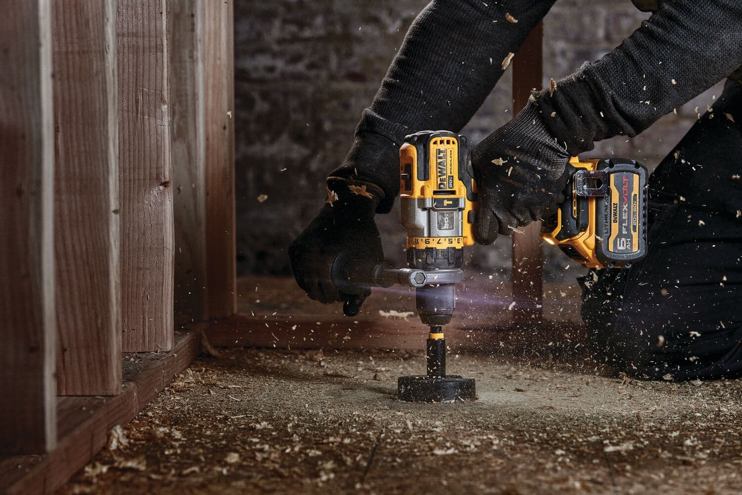 Una persona con guantes negros utiliza un taladro eléctrico amarillo y negro DEWALT para hacer un agujero en un suelo de madera, con virutas de madera volando alrededor.