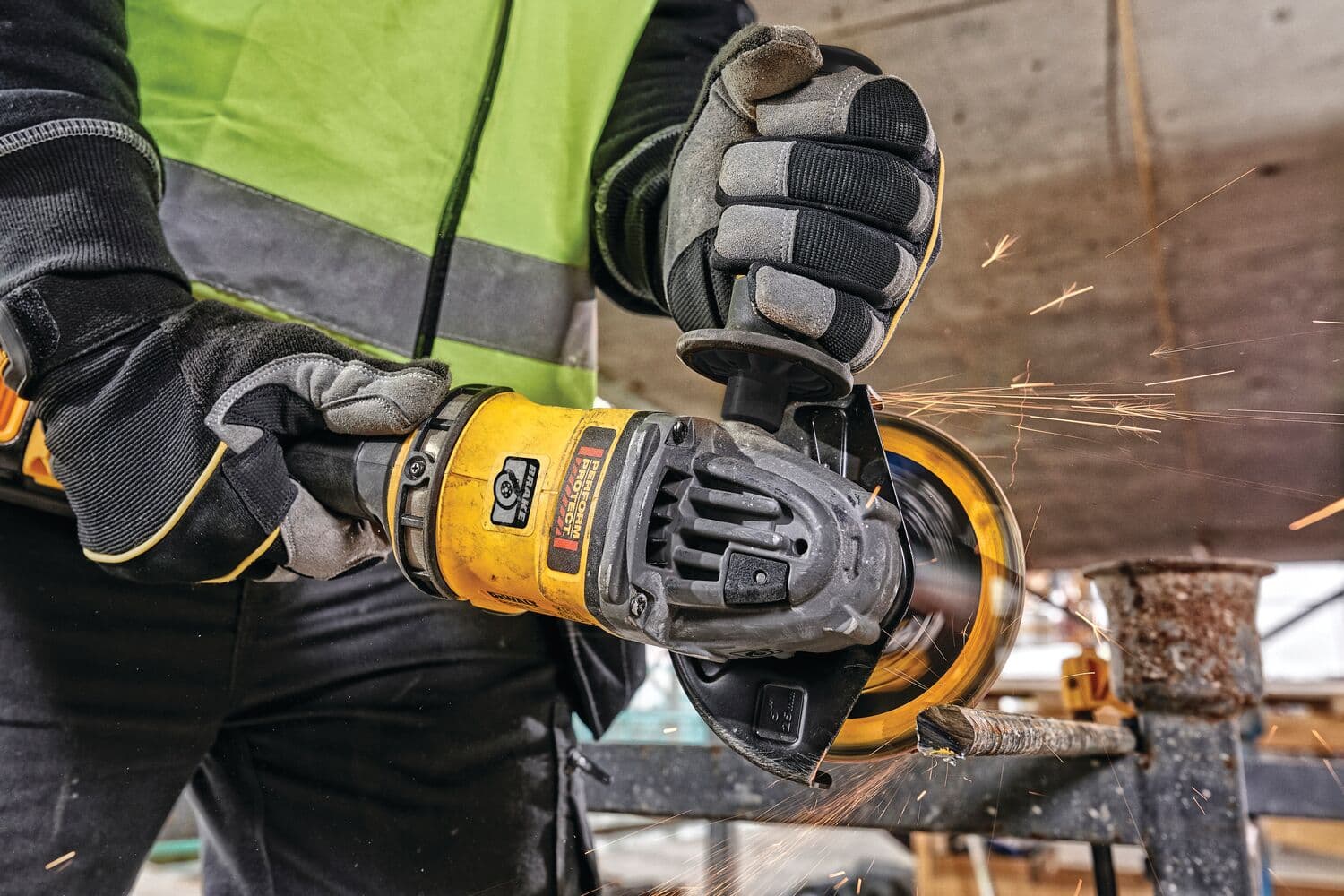 Una persona con guantes protectores y chaleco de seguridad utiliza una amoladora angular inalámbrica amarilla DEWALT para cortar metal, generando chispas en un entorno de construcción.
