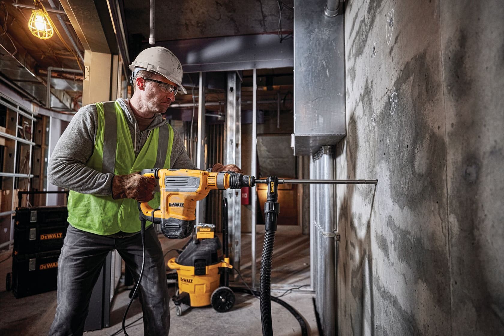 Una persona con equipo de seguridad y casco utiliza un martillo rotativo DEWALT D25614K sobre una pared de concreto en un sitio de construcción. Se pueden ver equipos y herramientas DEWALT en el fondo.