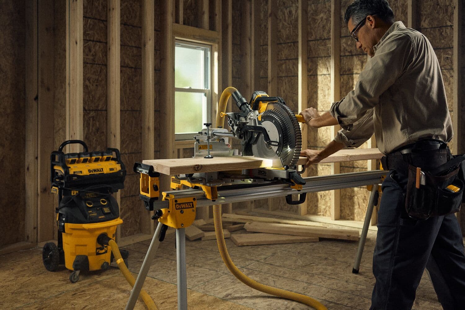 Una persona utiliza una sierra de inglete DEWALT montada en un soporte en un taller en construcción. En el fondo hay una aspiradora y una unidad de almacenamiento DEWALT amarillas. El espacio de trabajo está sin terminar, con estructuras de madera expuestas y una ventana.