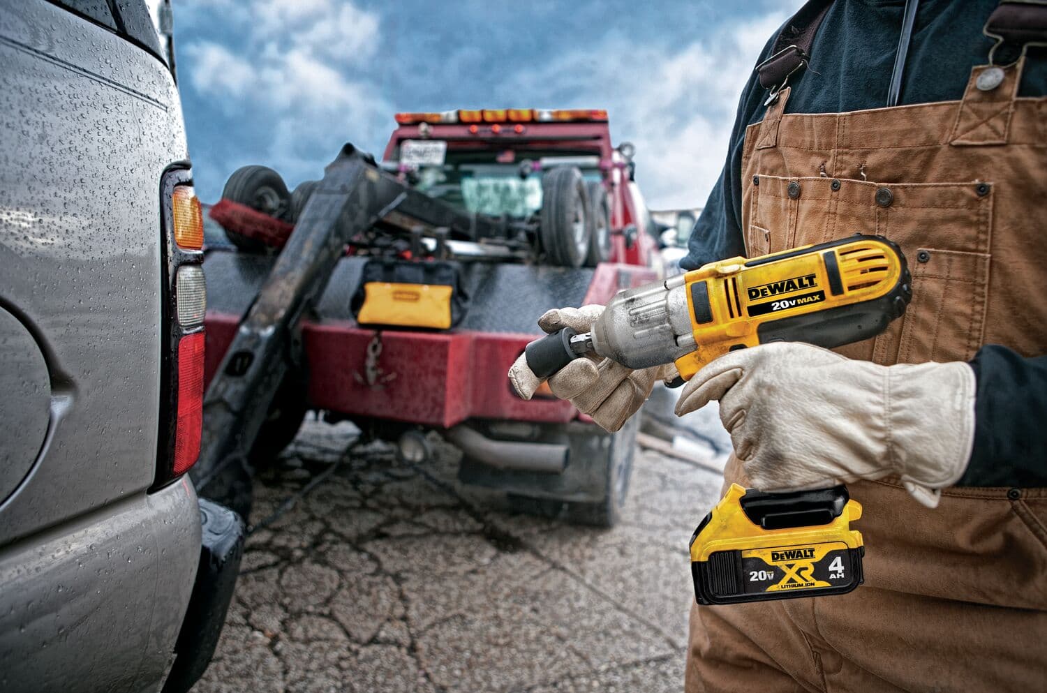 Una persona con guantes de trabajo y un overol marrón sostiene una llave de impacto inalámbrica DEWALT 20V MAX al aire libre, con una grúa y otro vehículo visibles en el fondo.