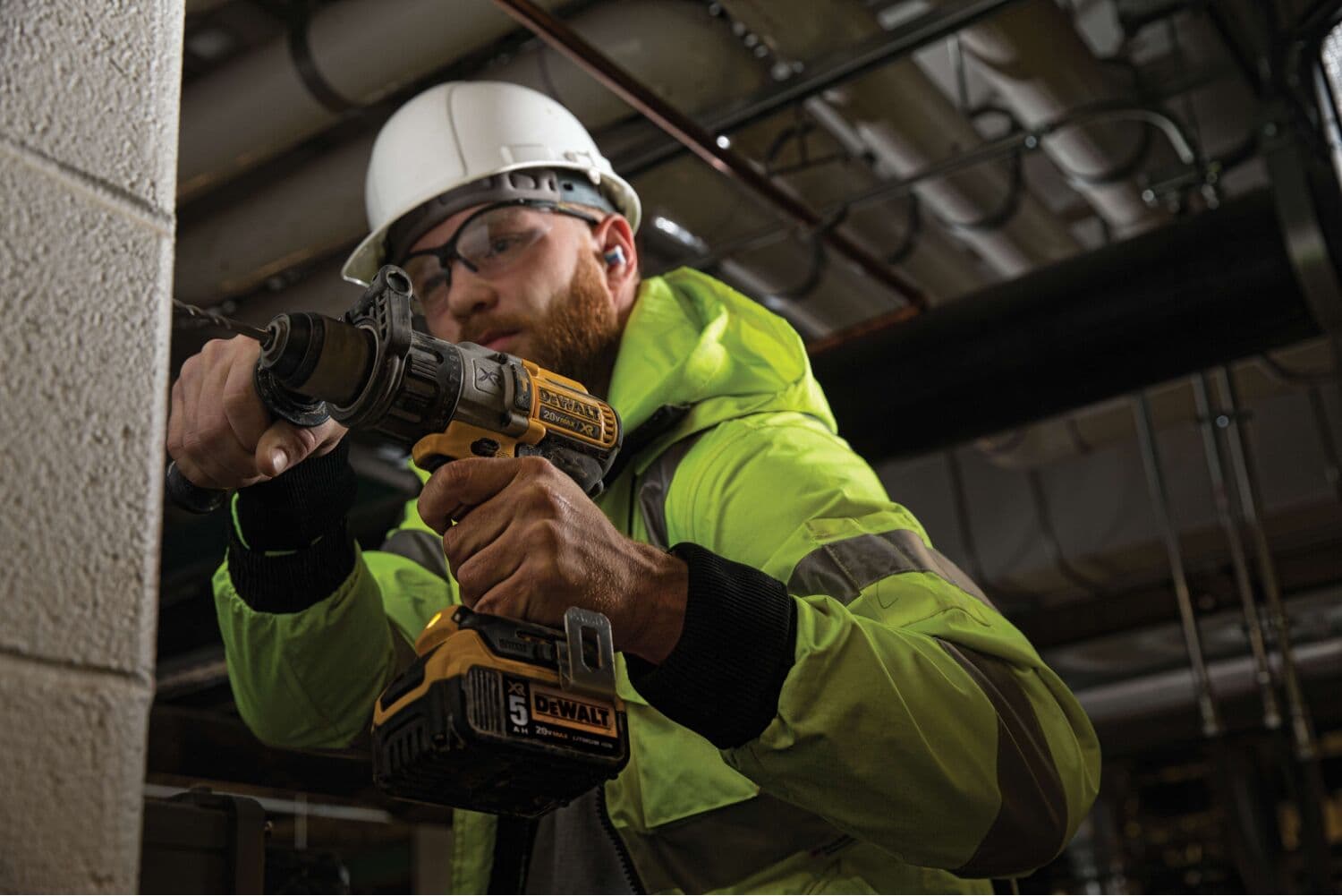 Una persona con chaqueta de alta visibilidad y casco de seguridad está utilizando un taladro atornillador inalámbrico DEWALT (modelo DCD996P2) para perforar una pared de concreto o ladrillo en un entorno industrial o de construcción. El producto y el agarre de la herramienta son claramente visibles.