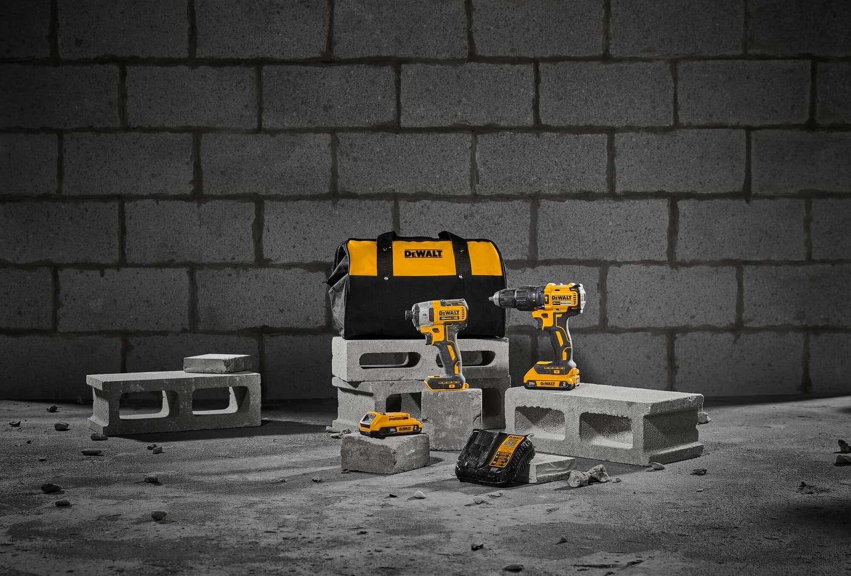 Un set de herramientas DEWALT que incluye una bolsa de transporte negra y amarilla, dos taladros inalámbricos, un paquete de batería y un cargador, exhibido sobre bloques de concreto frente a una pared de ladrillos.