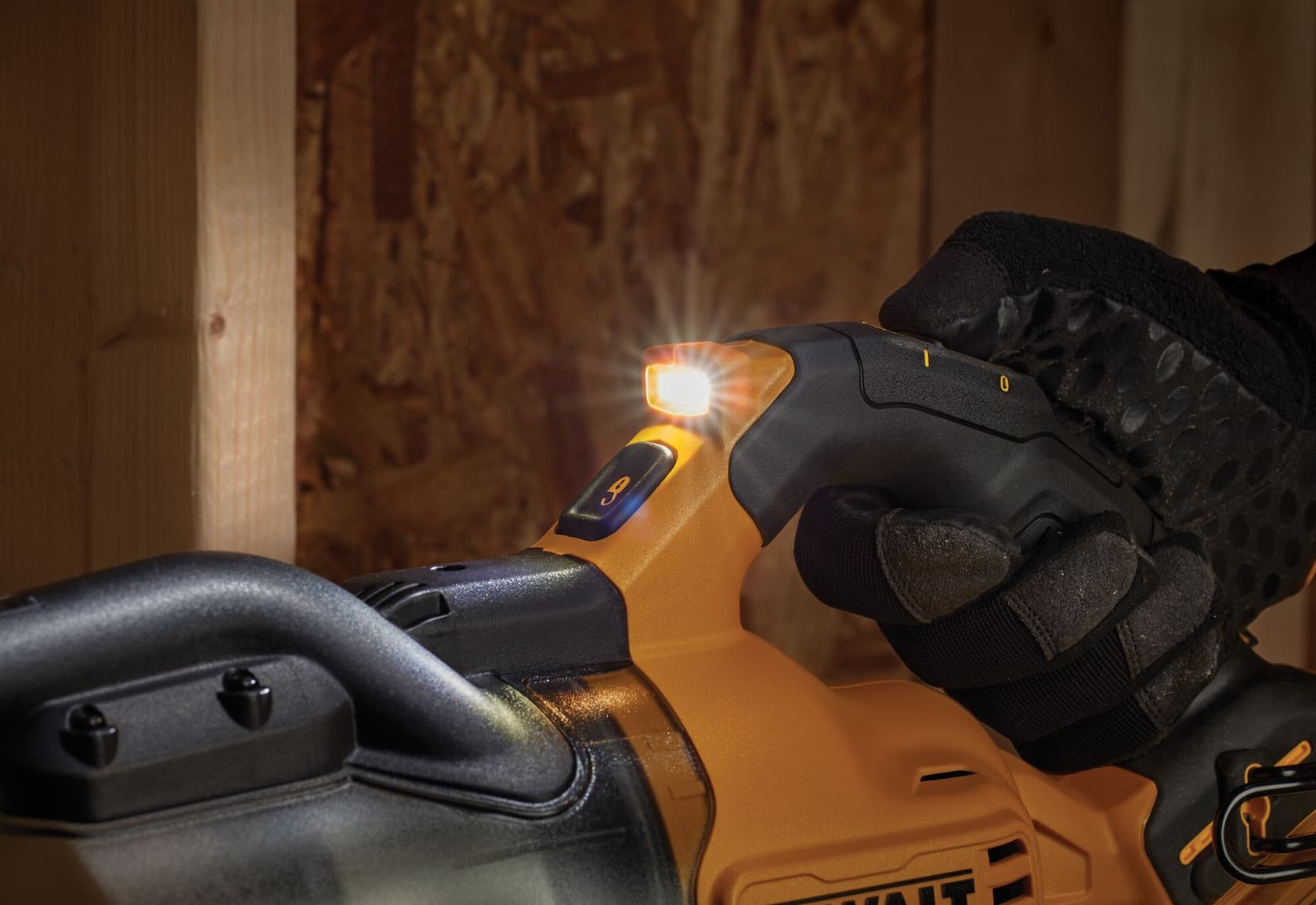 Primer plano de una mano con guante negro sujetando una herramienta eléctrica DEWALT con luz LED incorporada encendida, iluminando la zona cerca de superficies de madera.