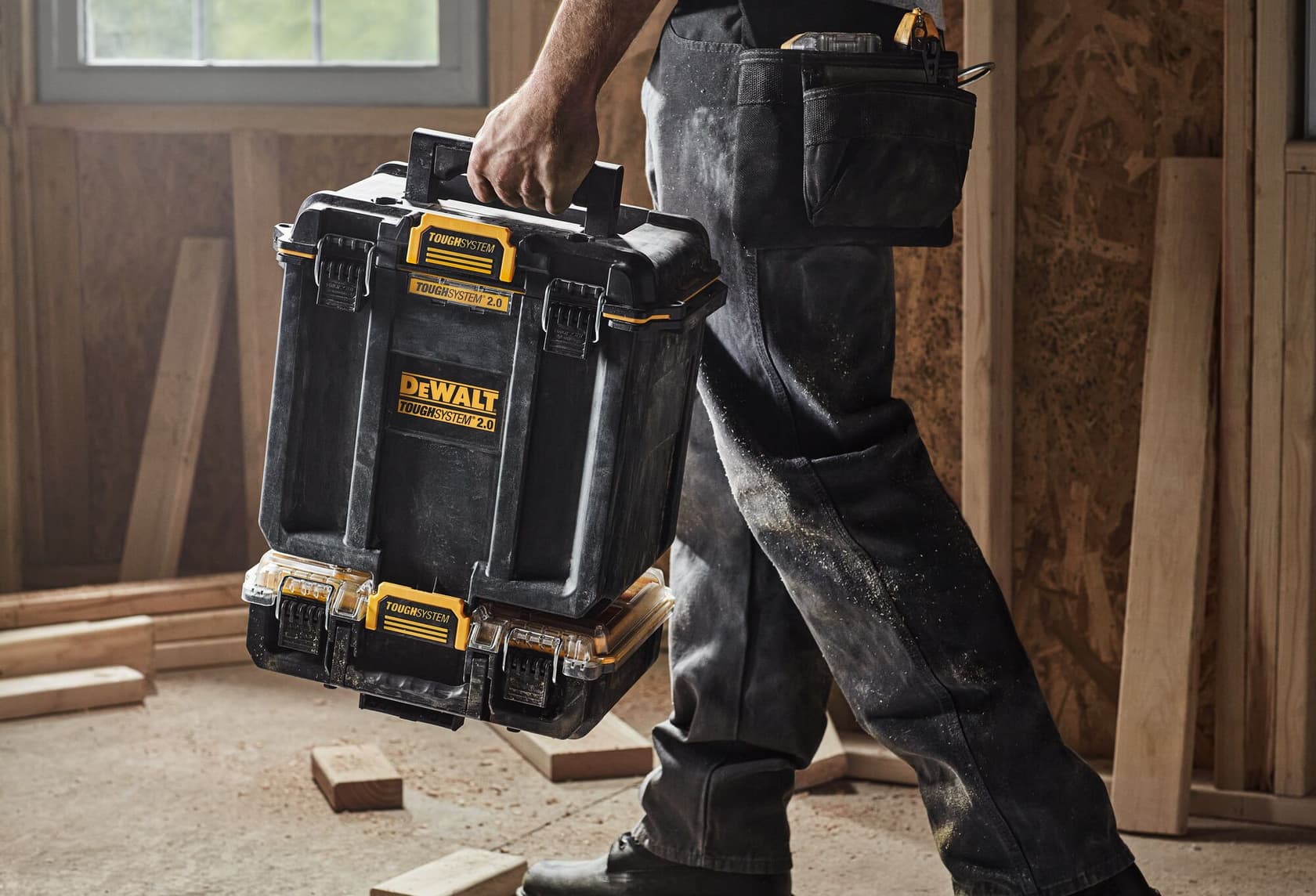 Una persona vestida con ropa de trabajo lleva una caja de almacenamiento DEWALT ToughSystem 2.0 dentro de un taller, con tablones de madera y aserrín visibles en el suelo.
