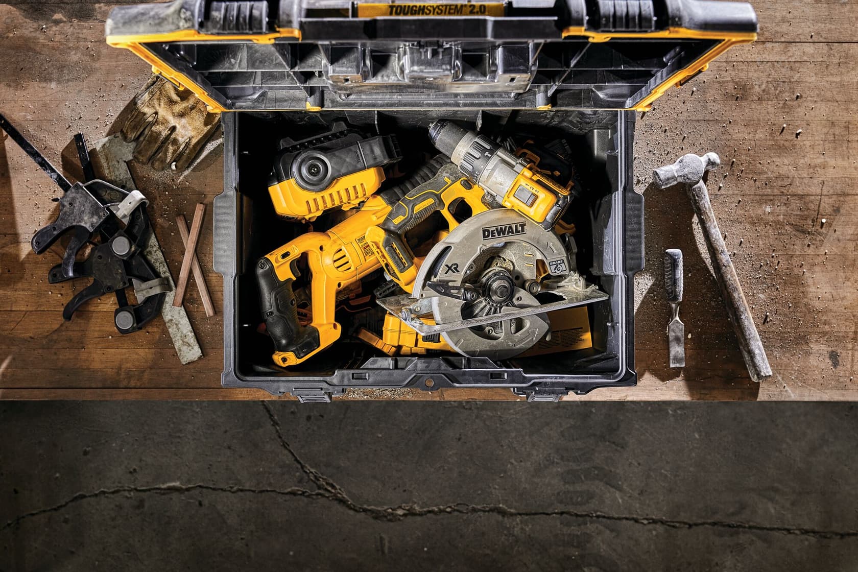 Una caja de herramientas DEWALT ToughSystem 2.0 abierta está sobre un banco de trabajo, contiene varias herramientas eléctricas amarillas y negras incluyendo una sierra circular y un taladro. Alrededor de la caja hay otras herramientas manuales como pinzas, un martillo, guantes y cinceles.