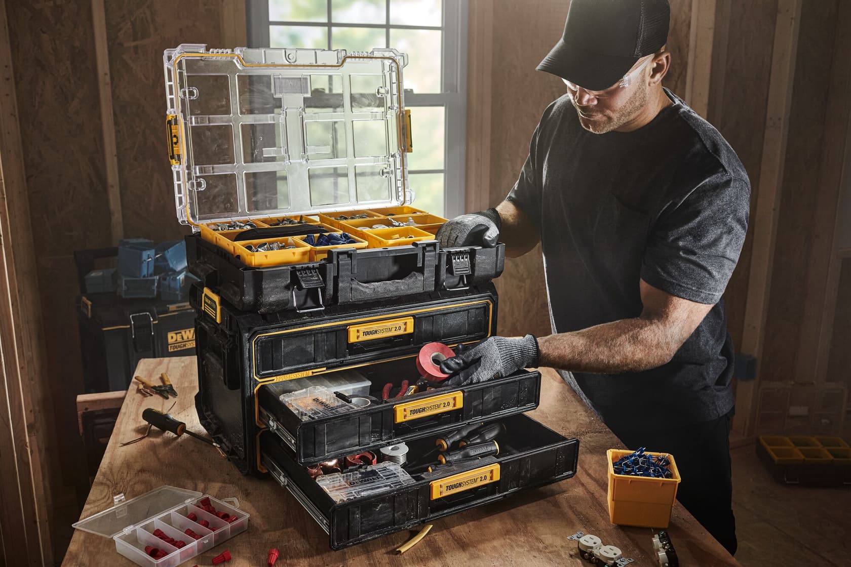 Una persona con guantes organiza herramientas y suministros en una caja de herramientas apilable DEWALT DWST08330 con tres cajones y un compartimento superior. La caja de herramientas está sobre una mesa de madera dentro de un taller.