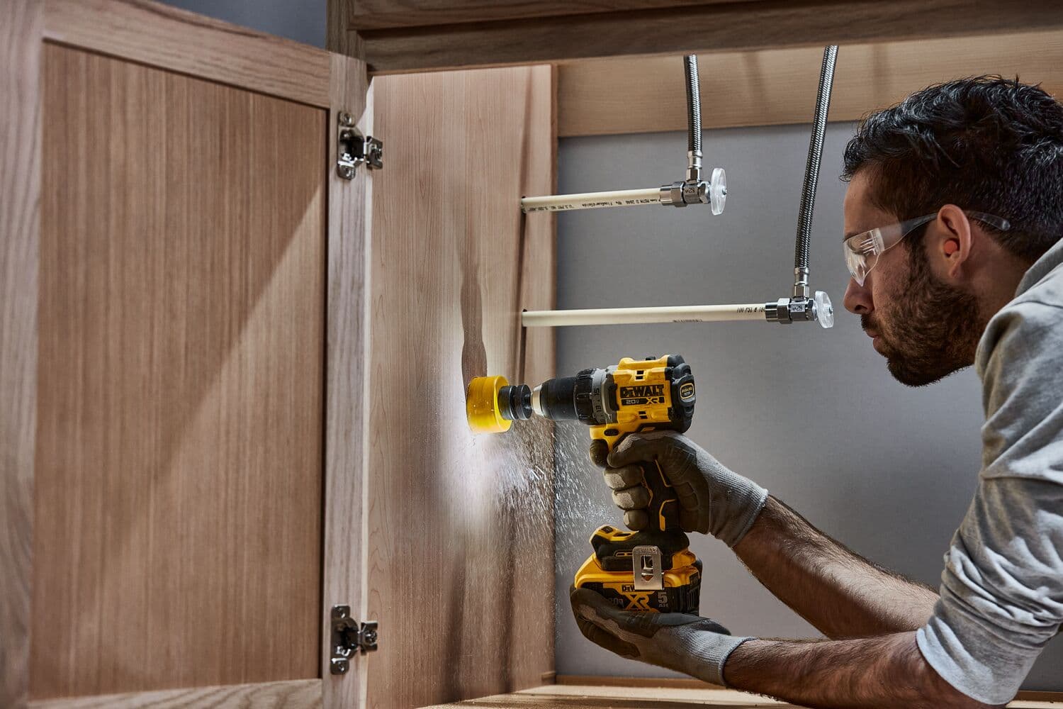 Una persona utiliza un taladro inalámbrico DEWALT dentro de un gabinete de madera, perforando un agujero cerca de tuberías de plomería. La cara de la persona está difuminada y lleva guantes de trabajo.