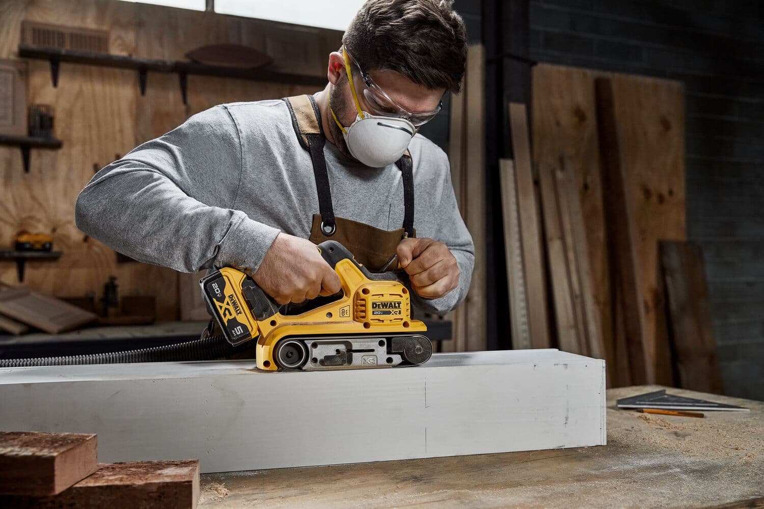 Una persona utiliza una lijadora de banda inalámbrica DEWALT (modelo DCW220B) sobre una tabla de madera en un taller. La persona lleva gafas de seguridad y un delantal. En el fondo se ven tablas de madera y herramientas de taller.