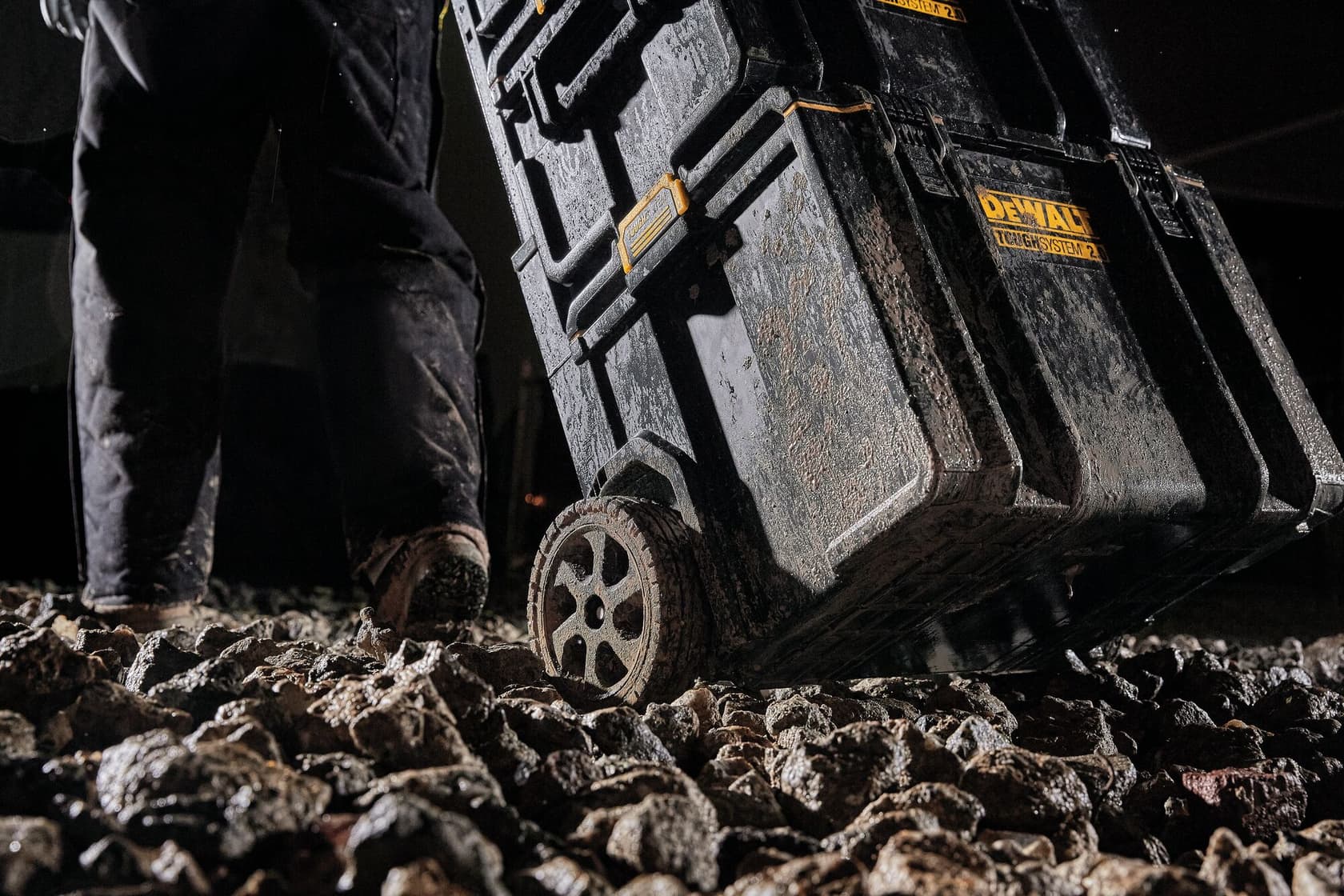 Una persona arrastra una caja de herramientas con ruedas DEWALT ToughSystem sobre un terreno rocoso y accidentado. La caja está cubierta de barro, lo que indica su uso en condiciones exteriores duras.