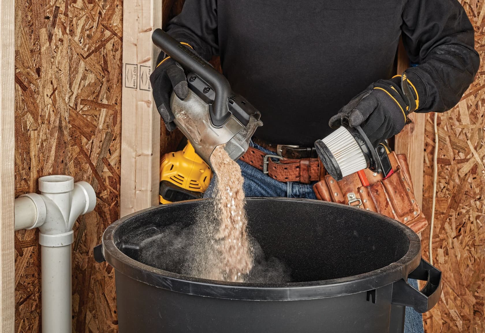 Una persona con guantes de trabajo vacía polvo y residuos de una aspiradora de mano DEWALT en un gran contenedor de basura negro. El filtro de la aspiradora está siendo sostenido en la otra mano. La escena se desarrolla en un taller con paredes de madera y fontanería visible.