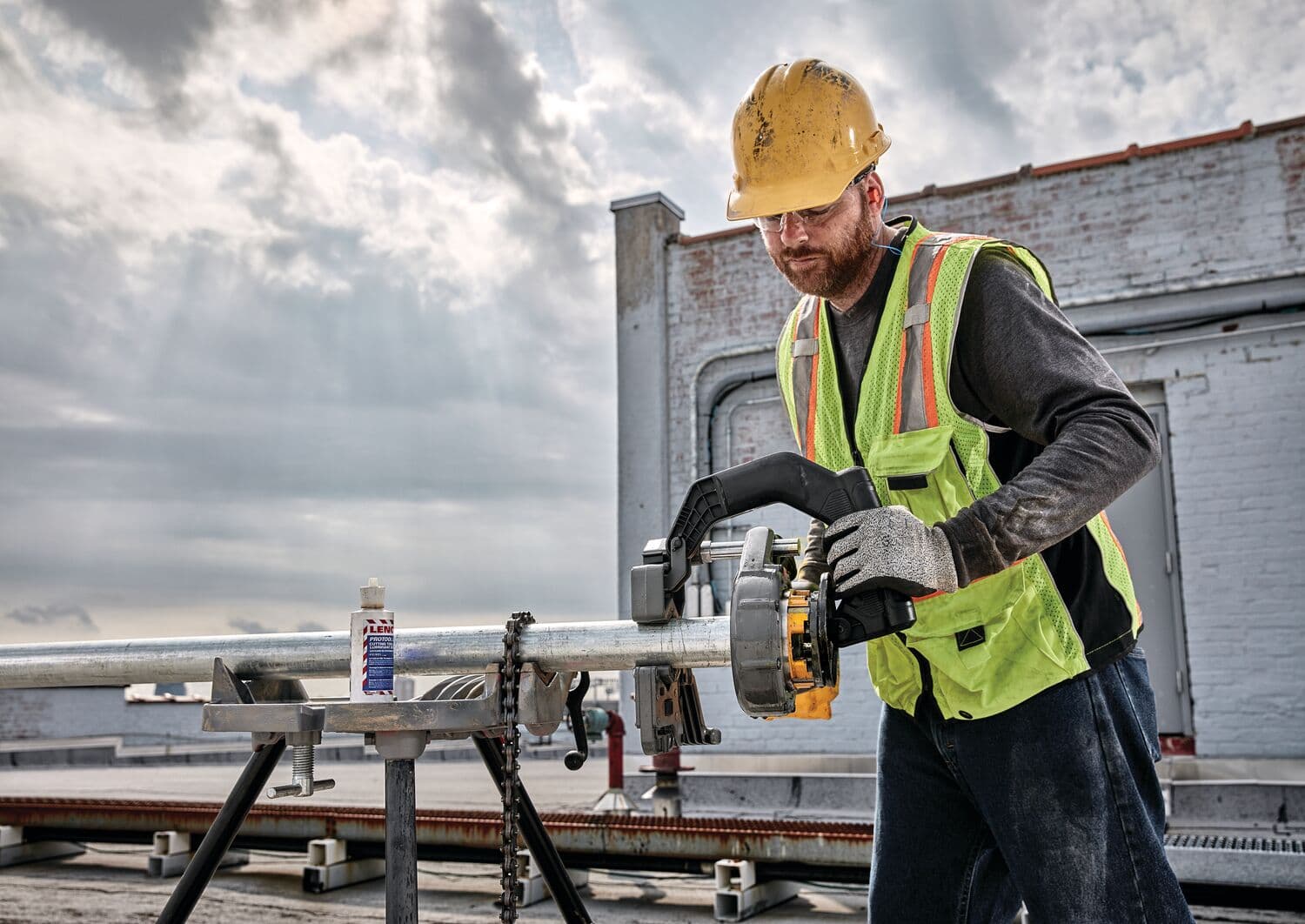 Un trabajador de construcción con casco amarillo, chaleco de seguridad y guantes utiliza una herramienta roscadora de tubos DEWALT sobre una tubería metálica en un sitio de trabajo al aire libre. El trabajador está usando la herramienta para roscar la tubería, que está asegurada en un soporte, con cielos nublados y un entorno industrial al fondo.