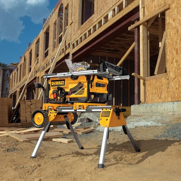 Se muestra una sierra de mesa portátil DEWALT con soporte en un sitio de construcción, con un edificio de madera parcialmente construido al fondo. La sierra y su soporte están colocados sobre suelo arenoso, con materiales de construcción como tablones de madera apilados cerca.