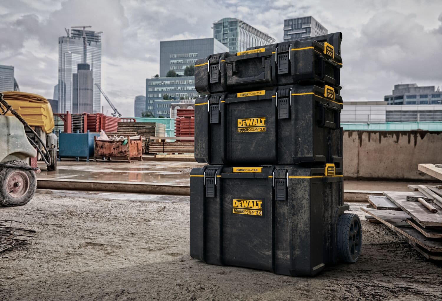 Pila de tres cajas de almacenamiento para herramientas DEWALT ToughSystem 2.0 negras con detalles amarillos, colocada en un sitio de construcción con edificios al fondo.