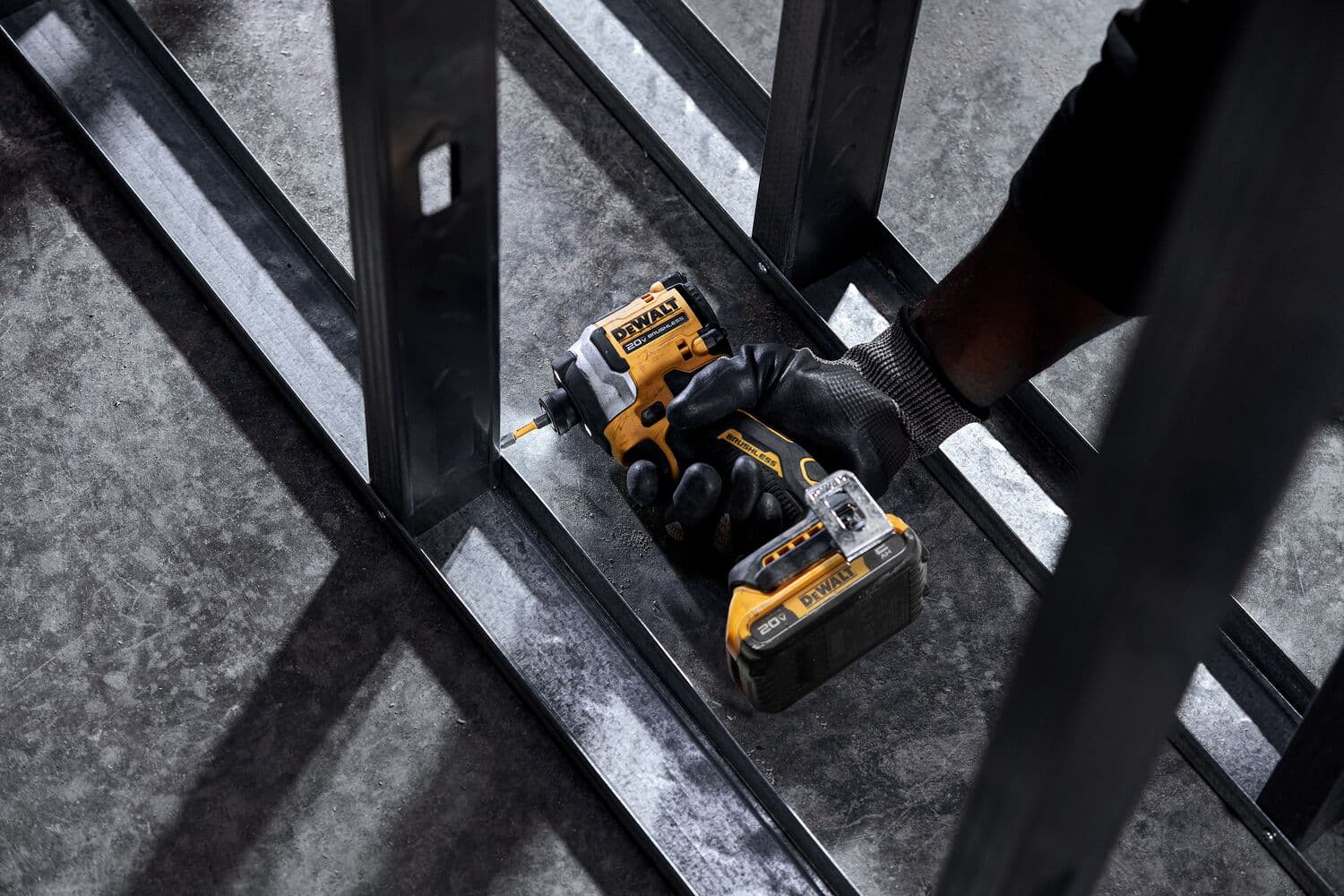 Una persona con guantes negros utiliza un destornillador de impacto inalámbrico DEWALT DCF850D2 para fijar tornillos en una estructura de marco de metal sobre un suelo de cemento.