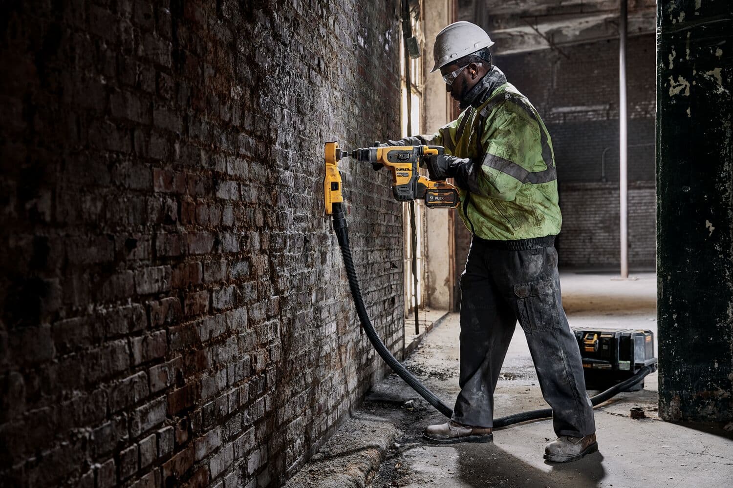 Un trabajador de la construcción con equipo de seguridad utiliza un taladro rotatorio inalámbrico DEWALT (DCH416B) con accesorio de extracción de polvo para perforar una pared de ladrillo dentro de un edificio industrial.