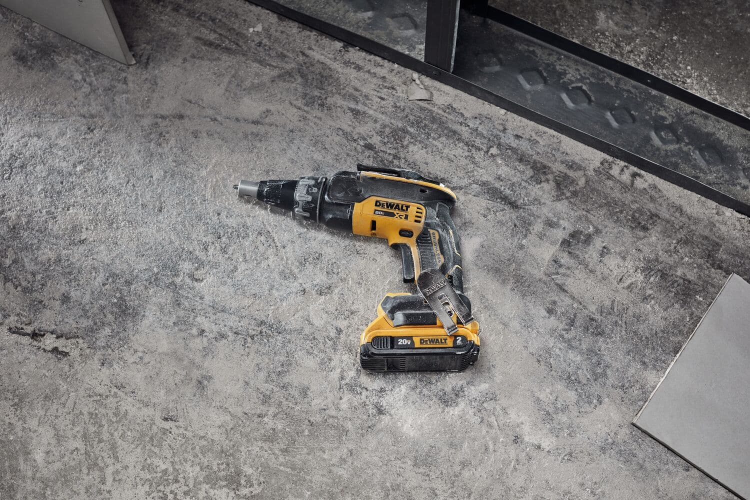 Un taladro inalámbrico DEWALT amarillo y negro está colocado sobre un suelo de concreto rugoso y polvoriento junto a unos materiales de construcción.