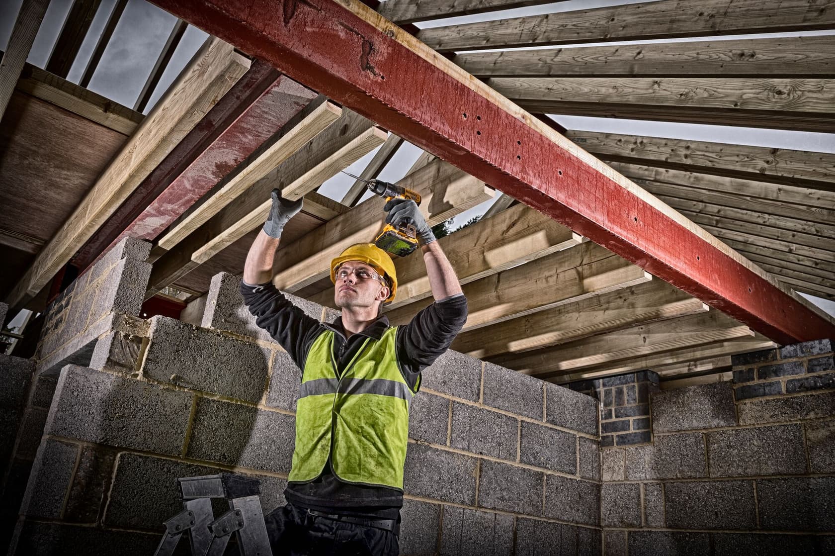 Un trabajador de la construcción con chaleco de seguridad verde y casco amarillo usa un taladro inalámbrico DEWALT para asegurar vigas de madera a una viga de acero roja en una estructura de mampostería parcialmente construida.