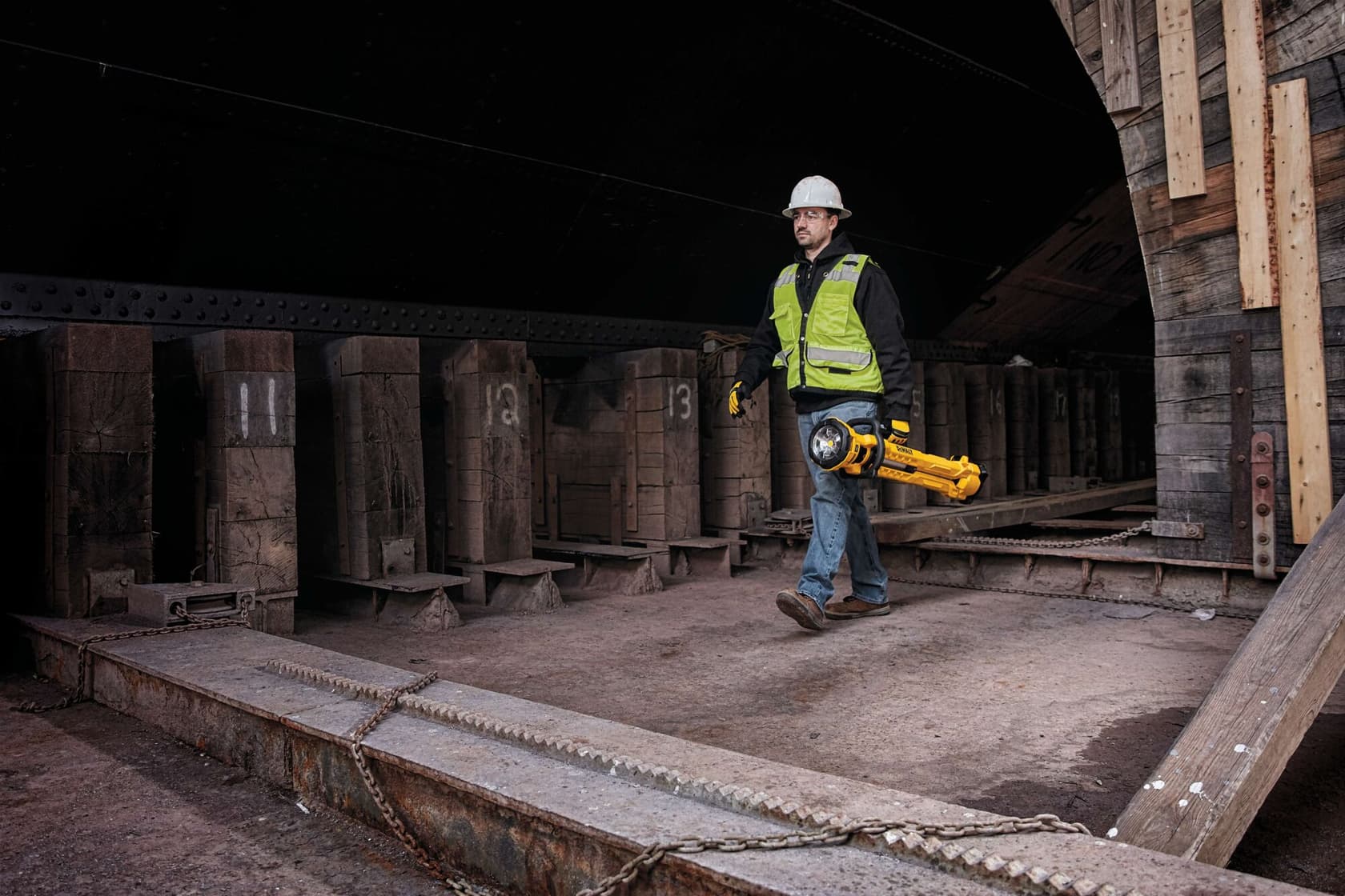 Un trabajador de construcción con chaleco de seguridad y casco camina en un entorno industrial, llevando una luz de trabajo LED portátil amarilla DEWALT con trípode (modelo DCL079R1). Al fondo se ven grandes vigas y estructuras de madera.