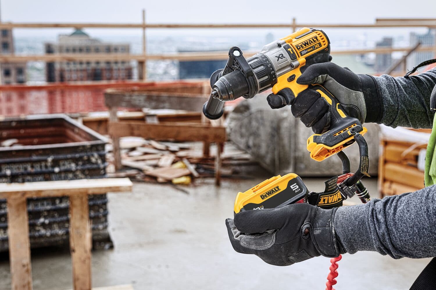 Una persona con guantes sostiene un taladro inalámbrico amarillo DEWALT con un paquete de baterías en un sitio de construcción al aire libre. Se pueden ver madera, concreto y materiales de construcción en el fondo.