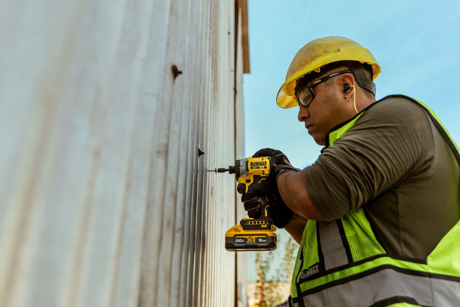 Trabajador usa atornillador DEWALT DCF860B en pared metálica, usando equipo de seguridad en exterior.