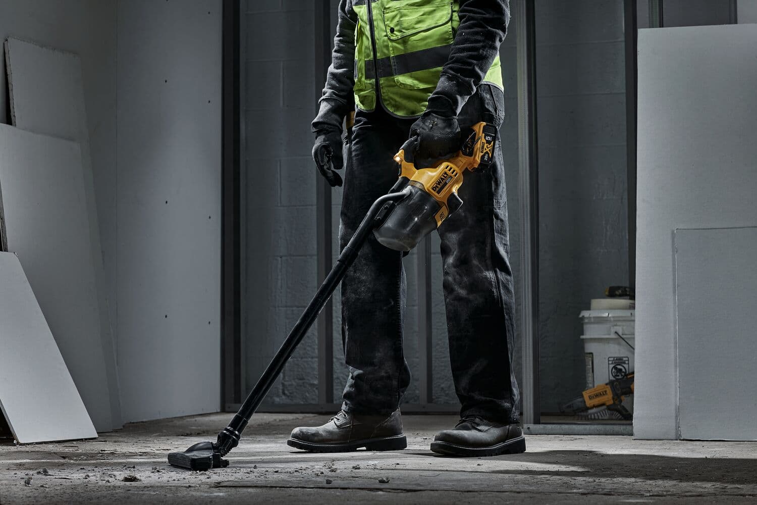 Una persona con chaleco de seguridad de alta visibilidad y guantes usa una aspiradora inalámbrica amarilla y negra DEWALT para limpiar polvo y residuos de un suelo de concreto en una habitación sin terminar.