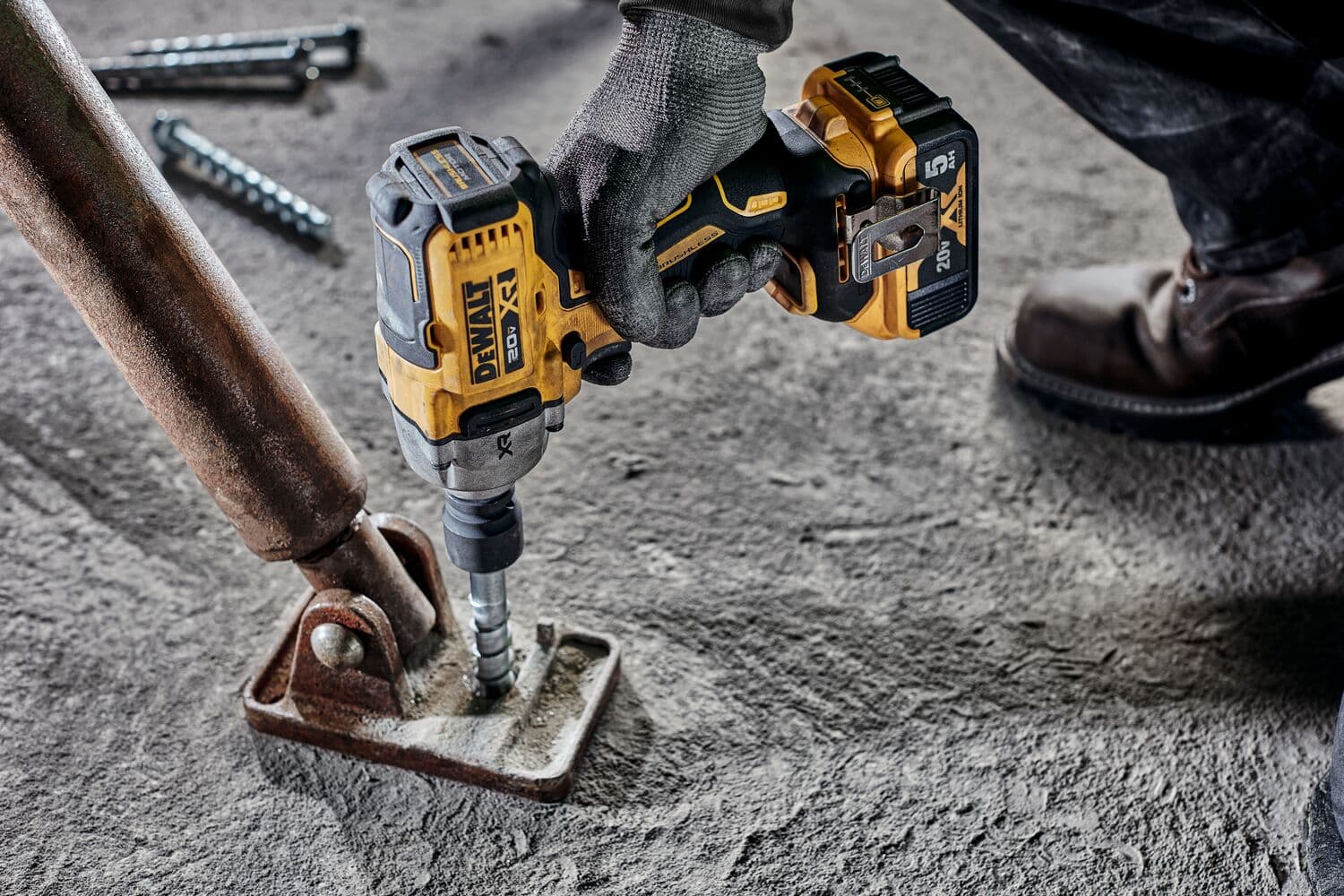 Una persona con guantes de trabajo utiliza una llave de impacto inalámbrica DEWALT para asegurar un perno de metal en un soporte metálico fijado al suelo de concreto.