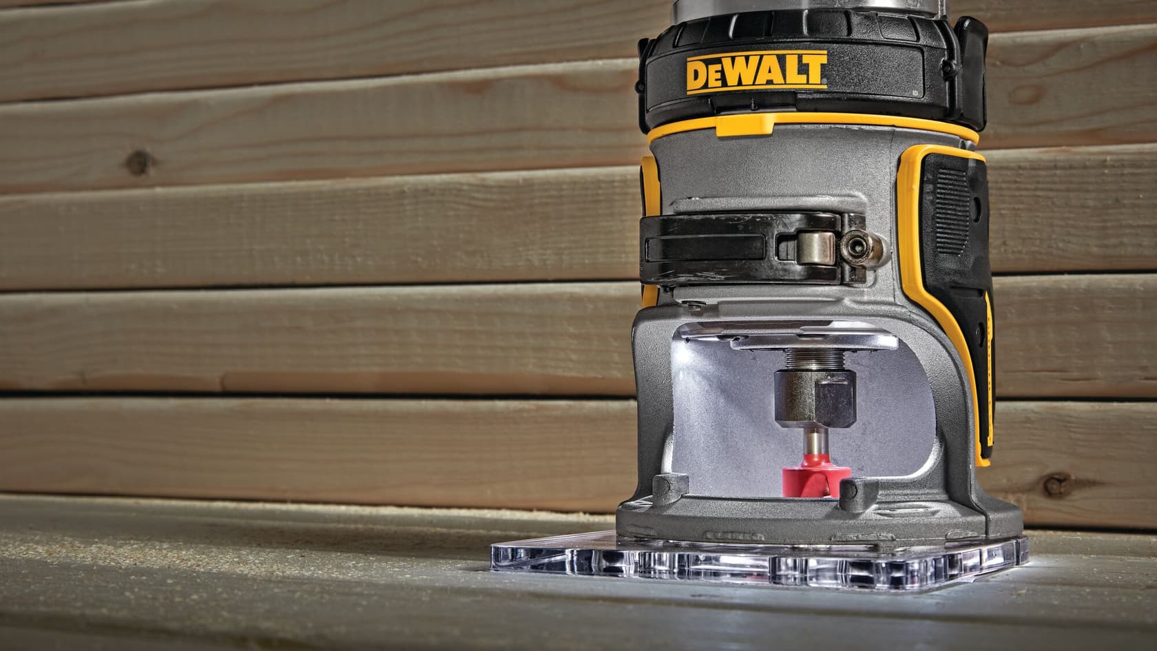Vista de primer plano de una fresadora compacta inalámbrica DEWALT, modelo DCW600B, colocada sobre una superficie de trabajo con una pared de madera al fondo.