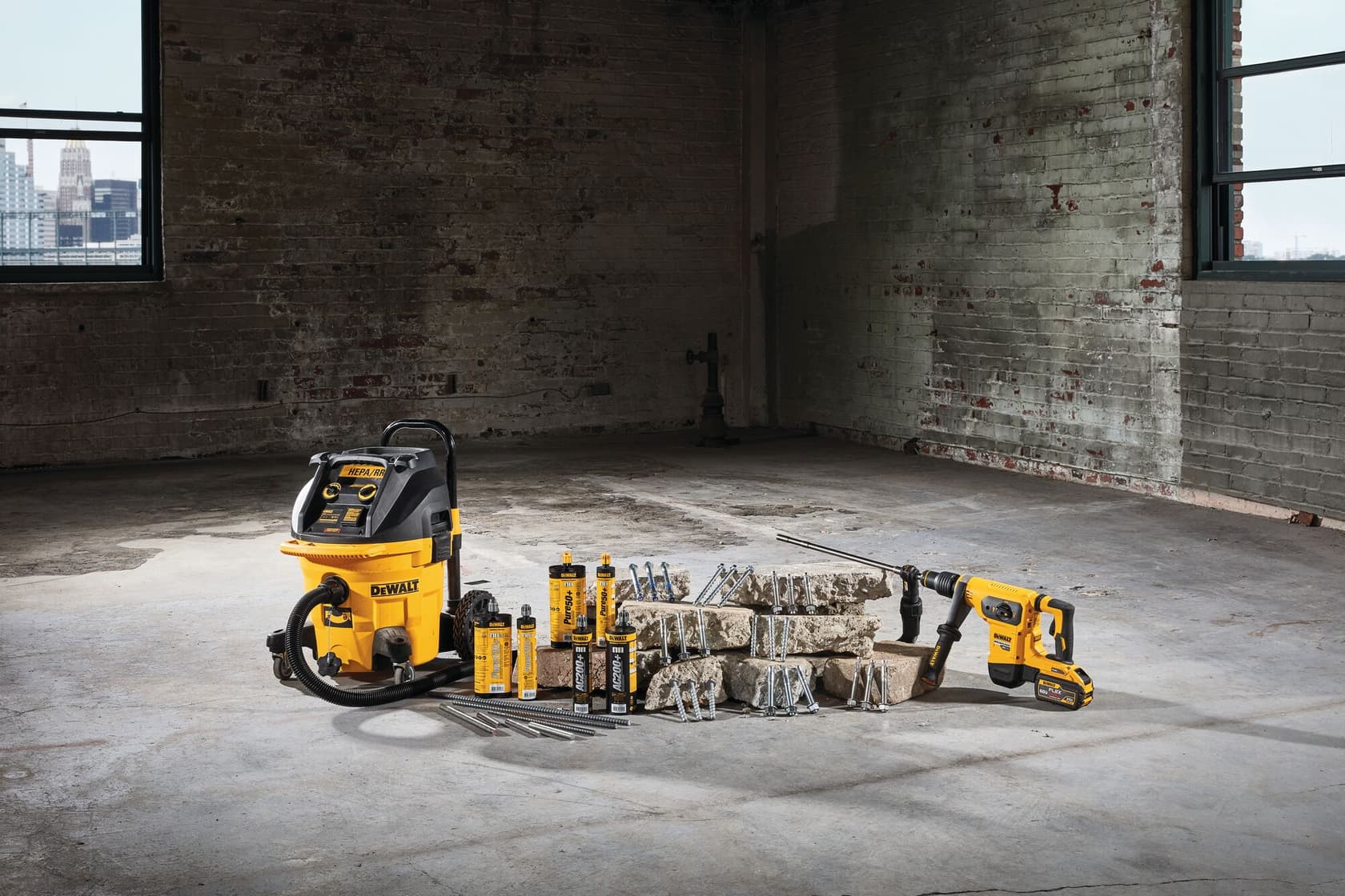 Una aspiradora industrial DEWALT, un taladro eléctrico y una colección de brocas y accesorios están exhibidos sobre un piso de concreto en un amplio almacén vacío con paredes de ladrillo visto y ventanas.