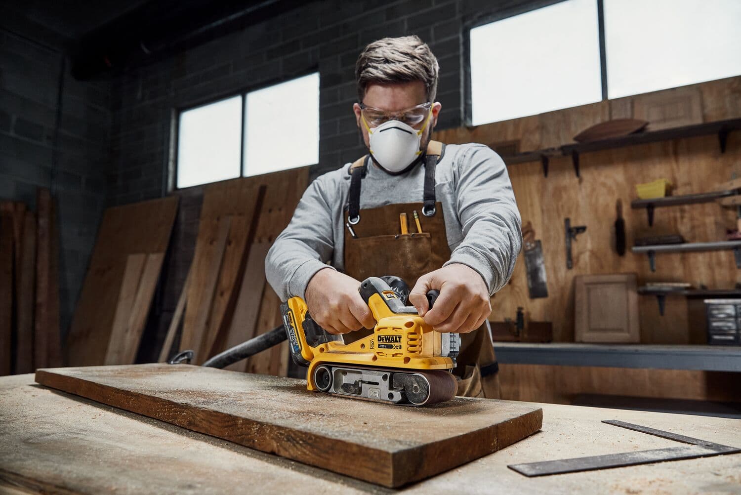 Una persona usando una lijadora de banda inalámbrica DEWALT DCW220B sobre una tabla de madera en un taller. El rostro de la persona está difuminado por privacidad.