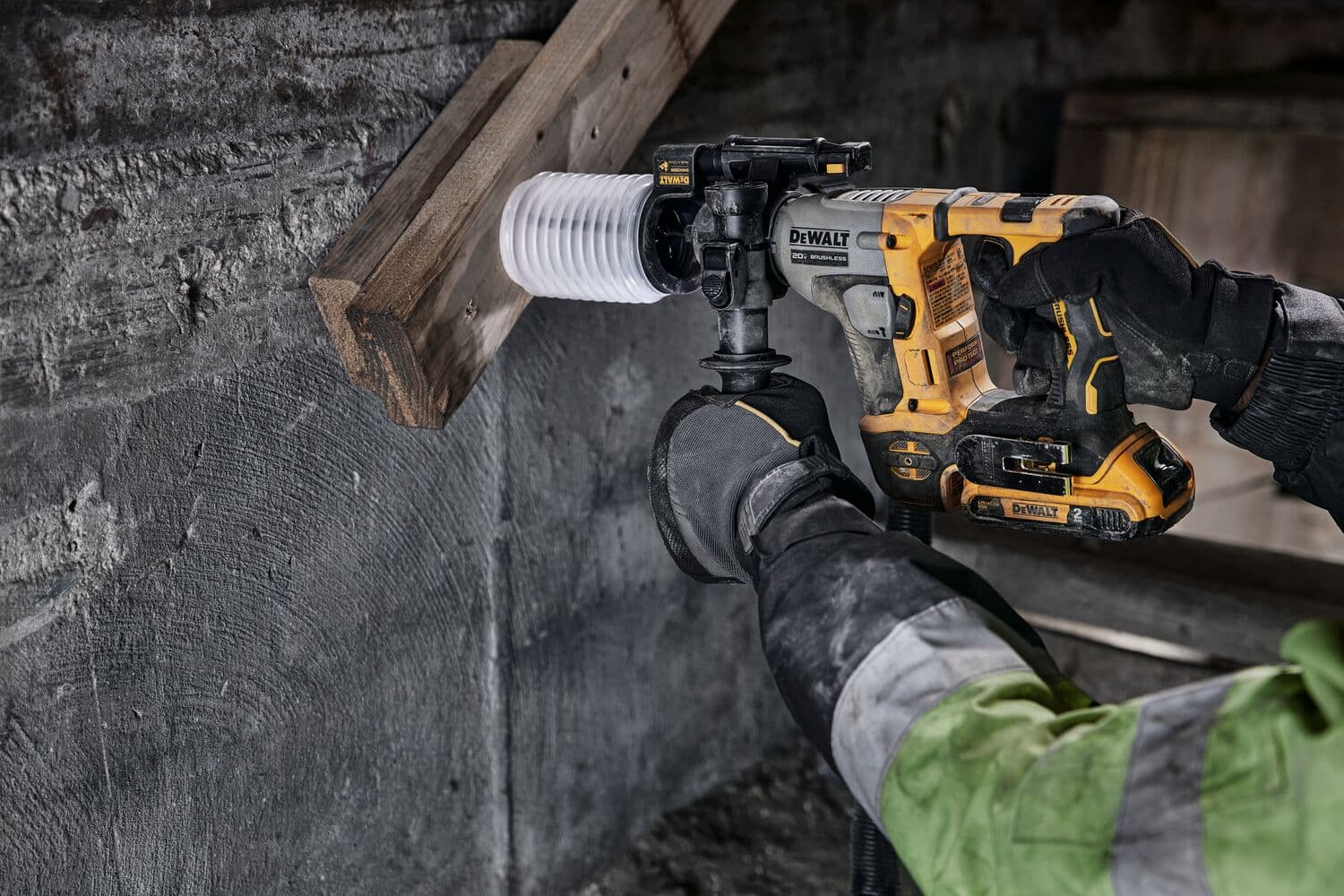 Una persona utiliza un taladro rotativo inalámbrico DEWALT para perforar un agujero a través de una viga de madera y una pared de concreto. El usuario lleva guantes y una chaqueta de trabajo.