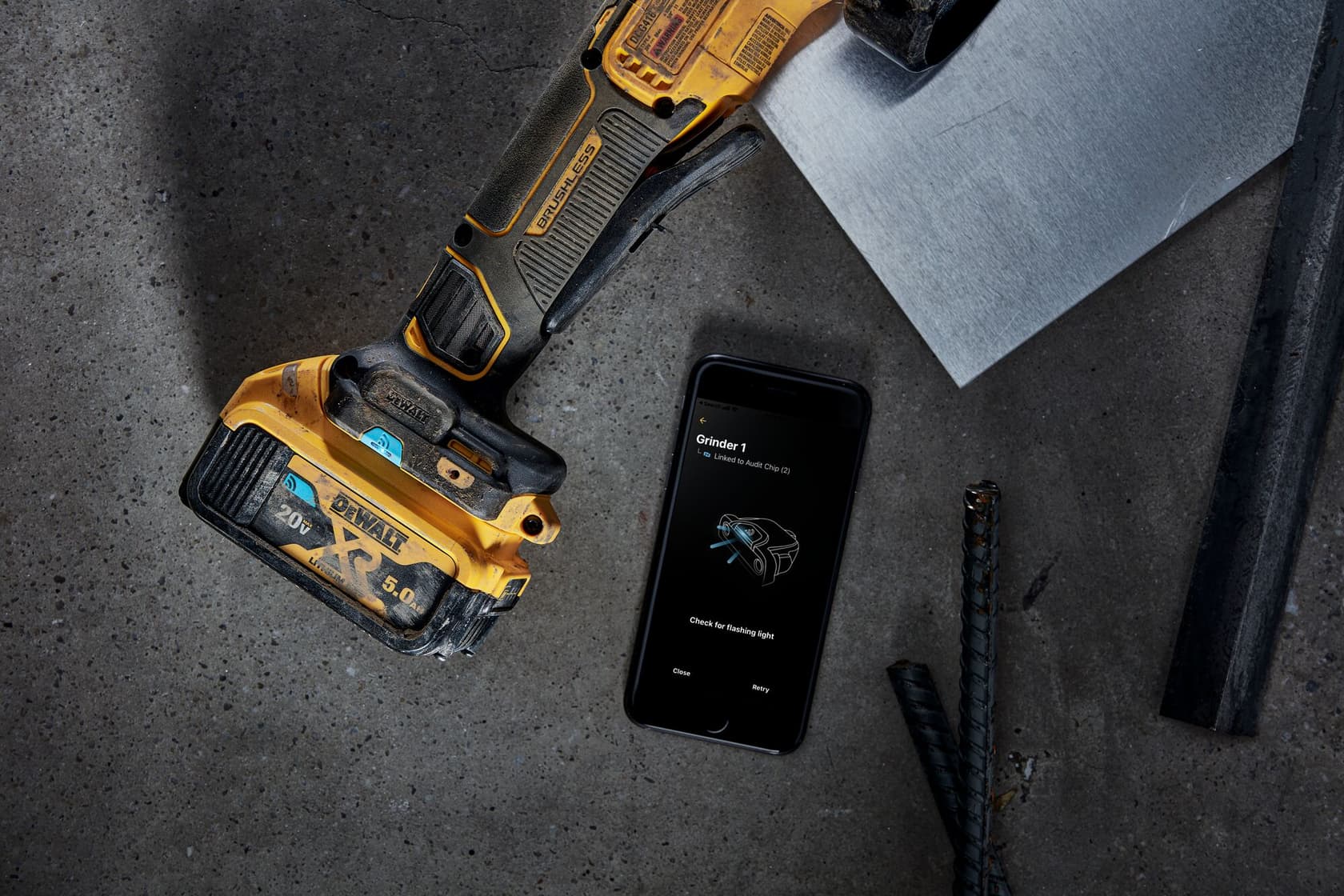 Una amoladora angular inalámbrica DEWALT está junto a un teléfono inteligente que muestra una aplicación relacionada con la amoladora sobre una superficie de concreto, con piezas de metal y varillas de refuerzo cercanas.