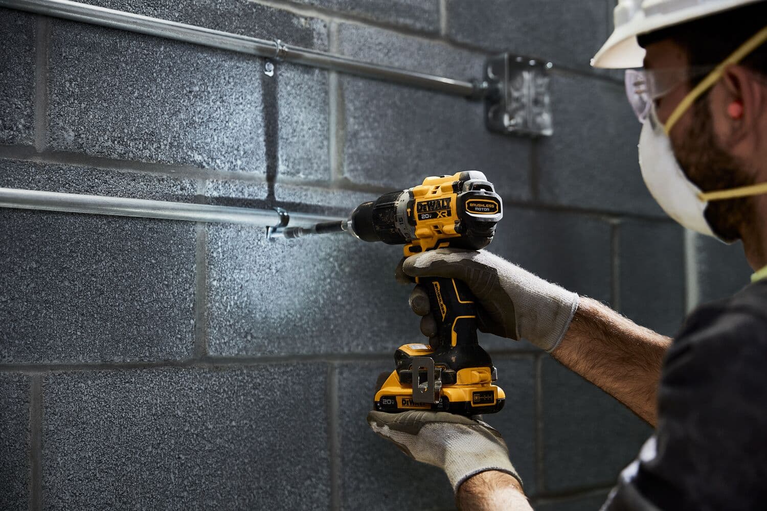 Una persona con guantes y casco de seguridad utiliza un taladro inalámbrico amarillo y negro DEWALT para trabajar en tuberías metálicas sujetas a una pared de bloques grises.