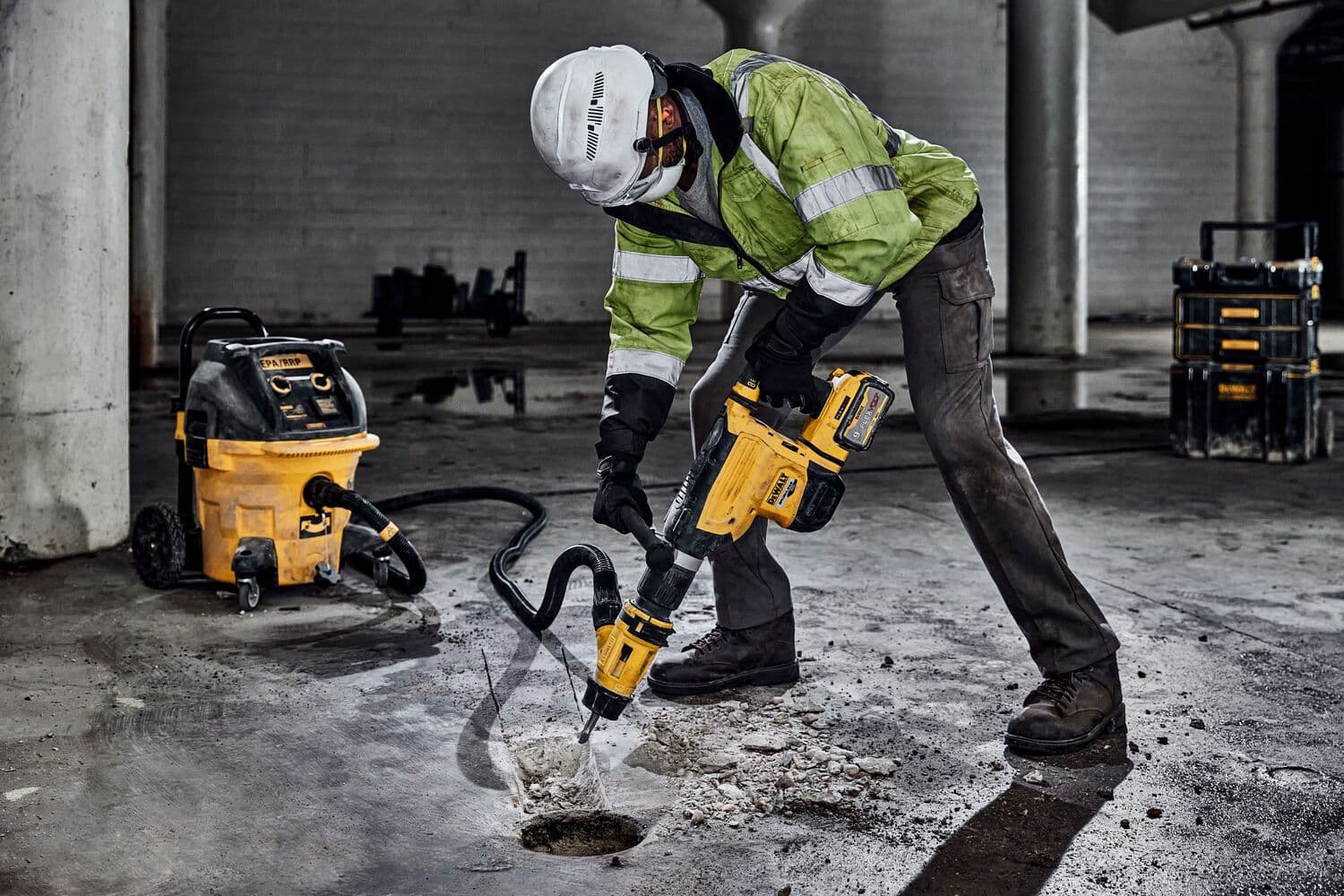 Un trabajador con equipo de seguridad opera un martillo perforador DEWALT para romper el concreto en una obra. Una aspiradora DEWALT está conectada a la herramienta para la extracción de polvo.