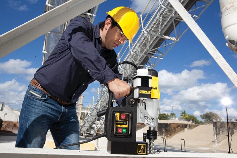 Una persona con casco amarillo y camisa azul opera un taladro magnético DEWALT DWE1622K sobre una superficie metálica en un sitio industrial al aire libre.