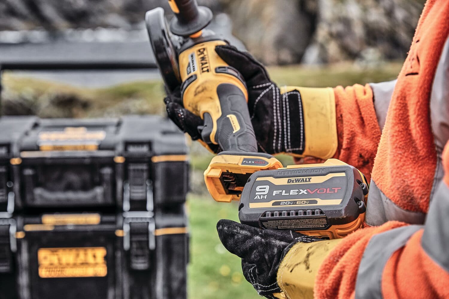 Una persona vestida con ropa de trabajo naranja y gris y guantes sostiene una batería DEWALT FLEXVOLT y una herramienta eléctrica. Se pueden ver cajas de almacenamiento negras de DEWALT en el fondo al aire libre.